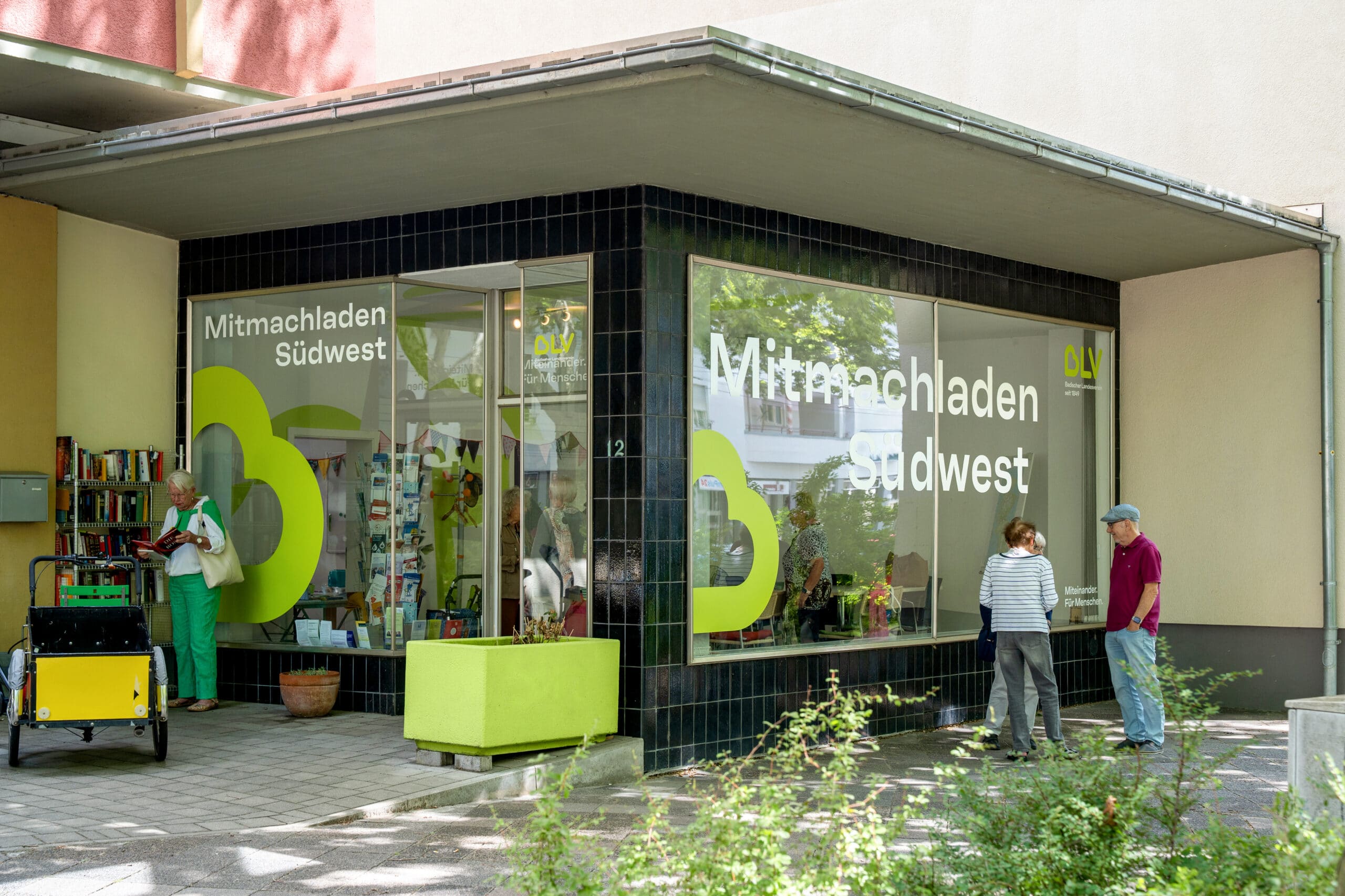 Der Mitmach-Laden Südwest von der Straße aus betrachtet. Eine Frau steht vor einem Bücherregal neben dem Eingang und blättert durch die Bücher. Drei weitere Personen stehen auf der anderen Seite und unterhalten sich.