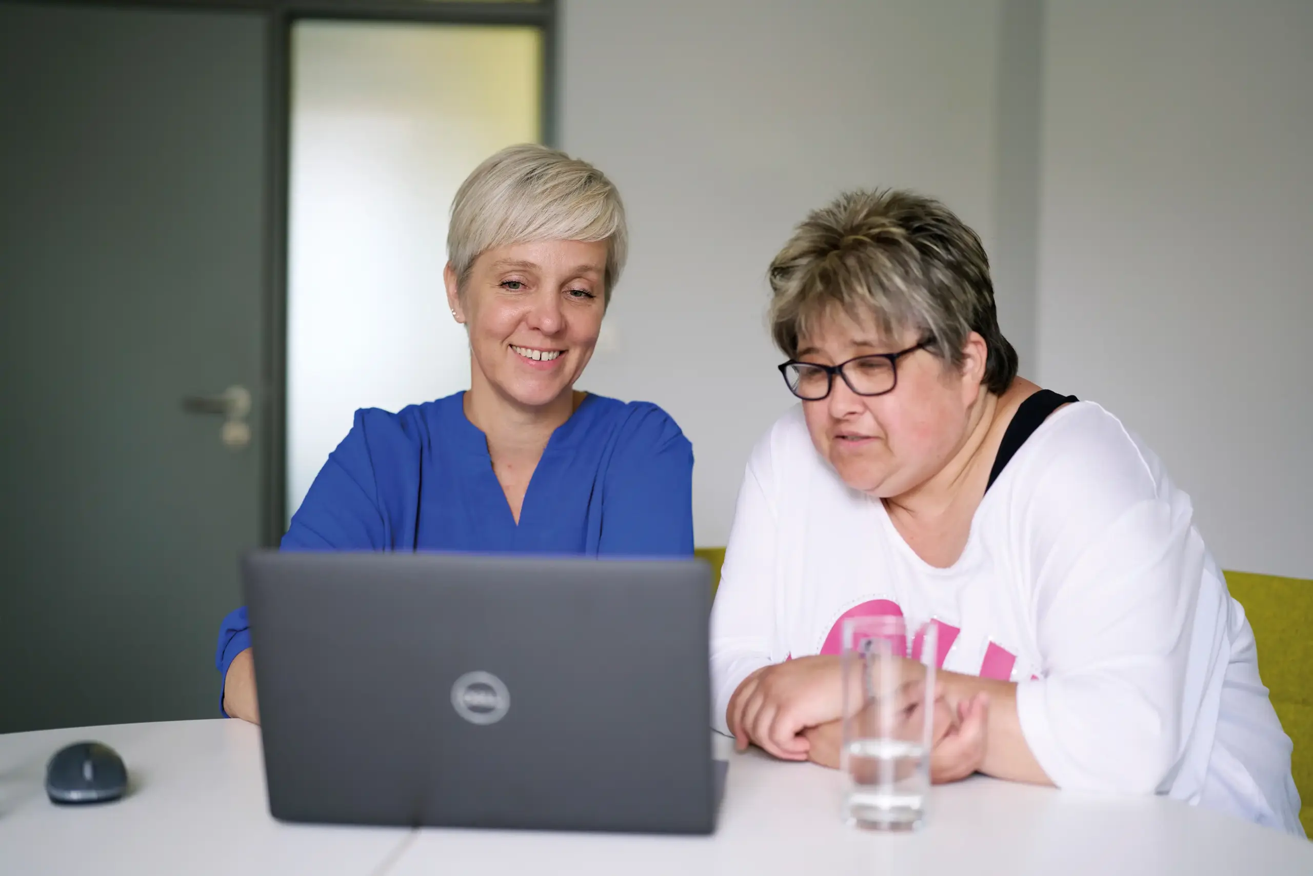 Zwei Frau sitzen an einem Tisch und schauen auf den Bildschirm eines Laptops. Sie lachen.