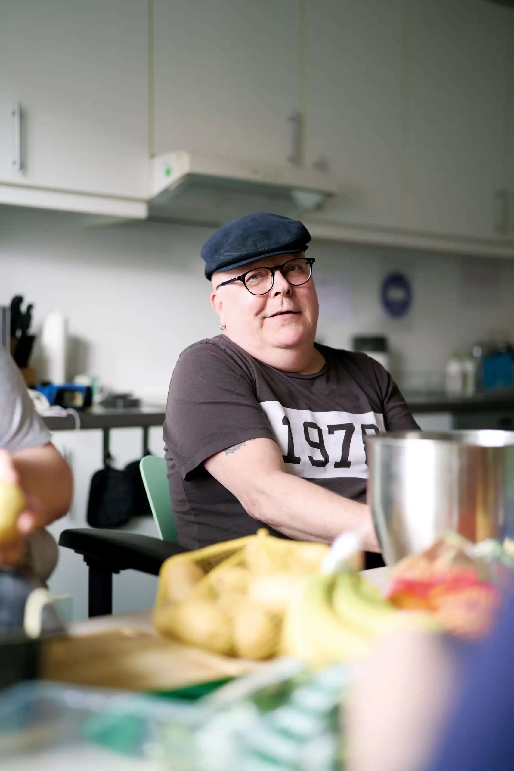 Quelle: BLV Ein Mann sitzt an einem Tisch. Er hat eine schwarze Schiebermütze auf, trägt eine Brille und ein braunes T-Shirt. Vor ihm auf dem Tisch liegt diverses Gemüse. Hinter ihm kann man vage eine Küche erkennen.