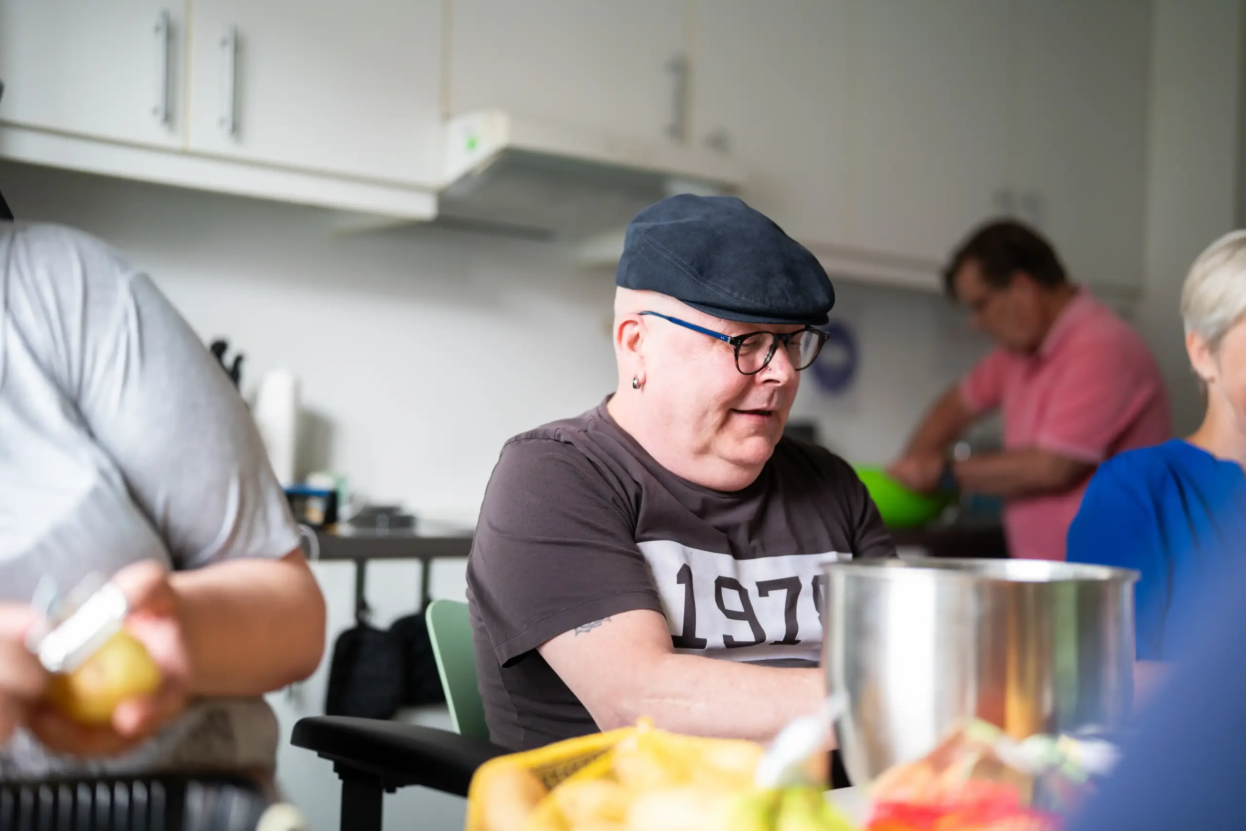 Ein Mann mit Schiebermütze sitzt an einem Tisch. Weitere Menschen sitzen rechts und links neben ihm. Sie schneiden Obst und Gemüse. Im Hintergrund kann man eine dritte Person sehen.