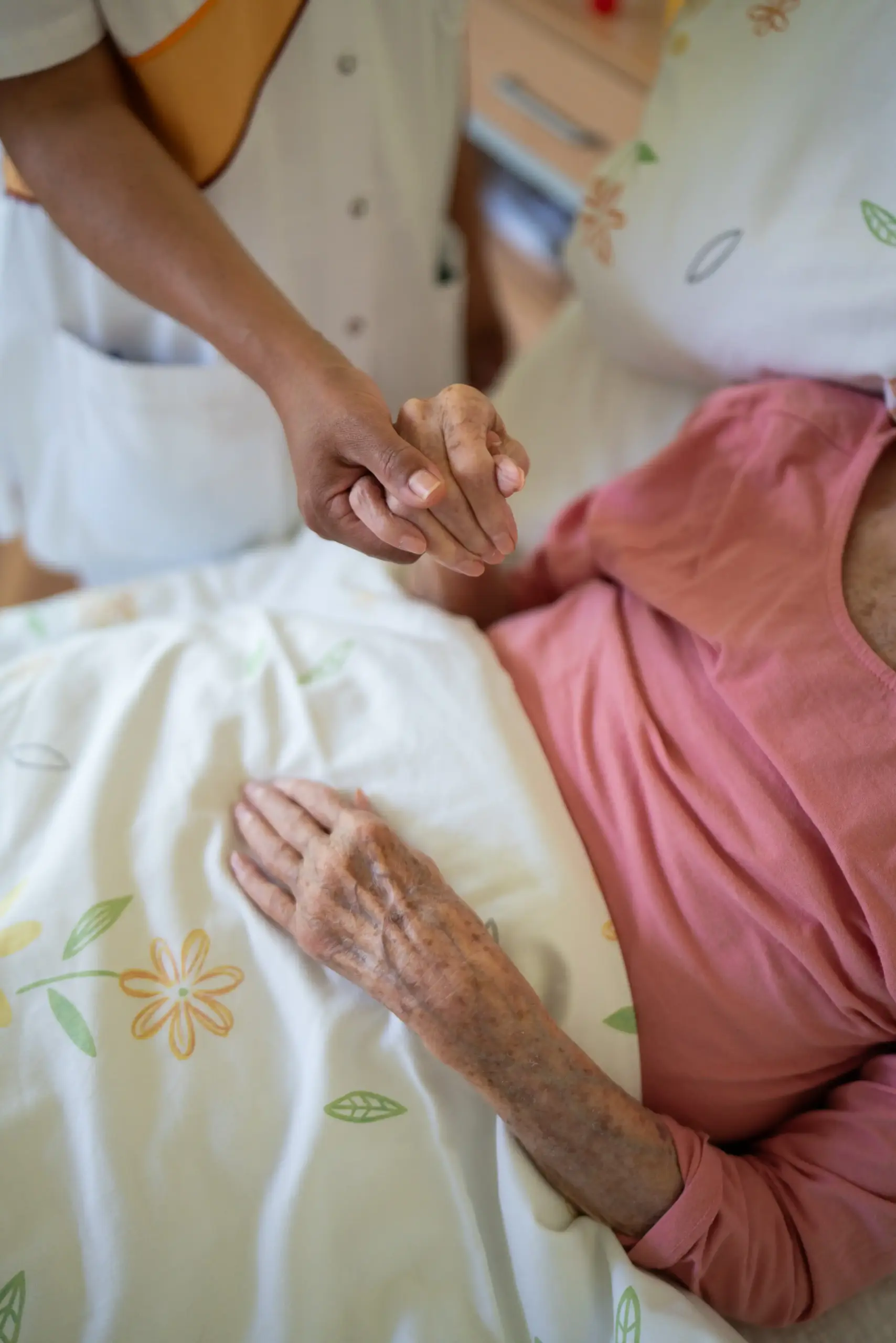 Eine junge Pflegerin hält die Hand einer älteren Frau, die im Bett liegt.