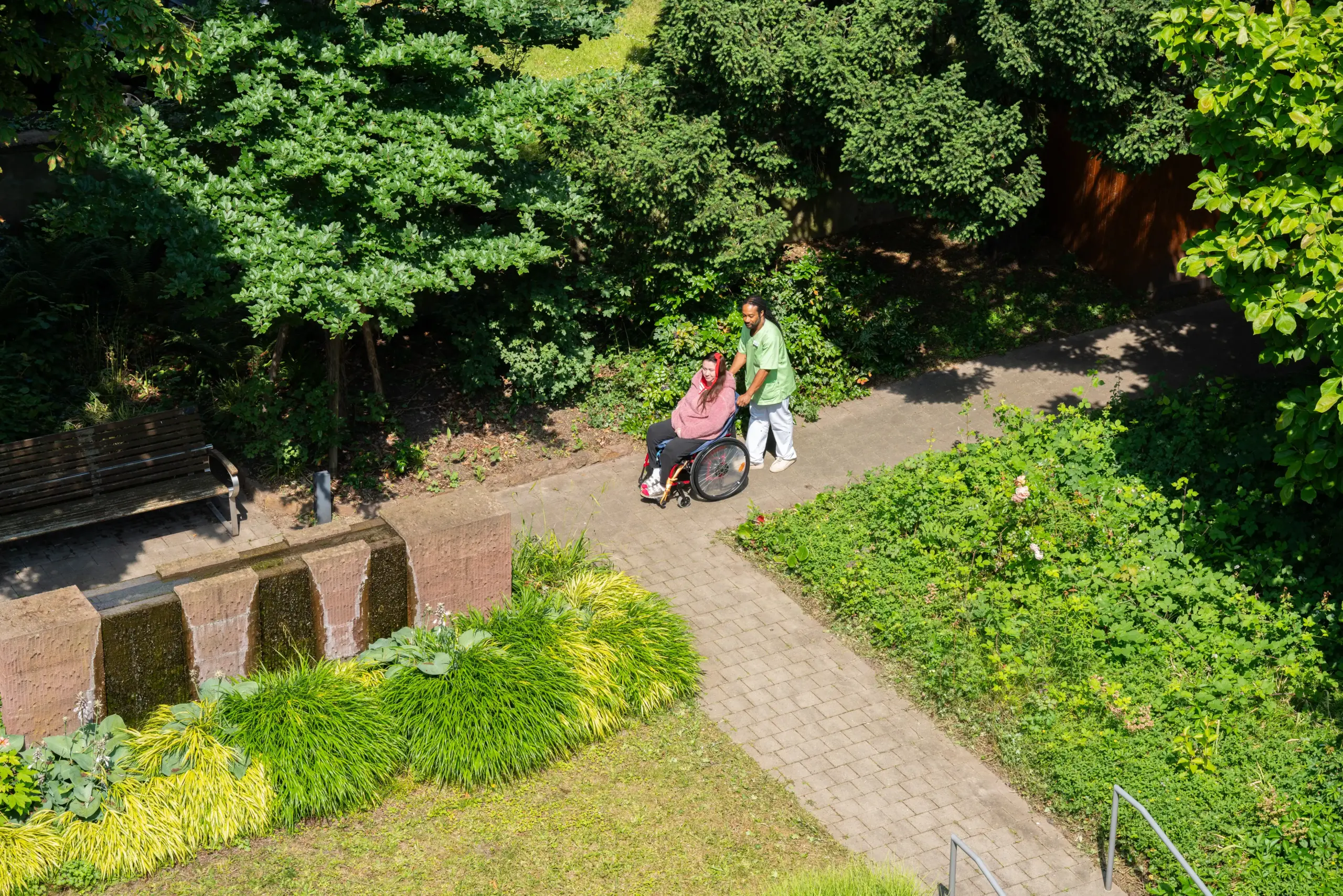 Ein Mann in grüner Pflegekleidung schiebt eine jüngere Frau in einem Rollstuhl im Garten des Friedensheims.