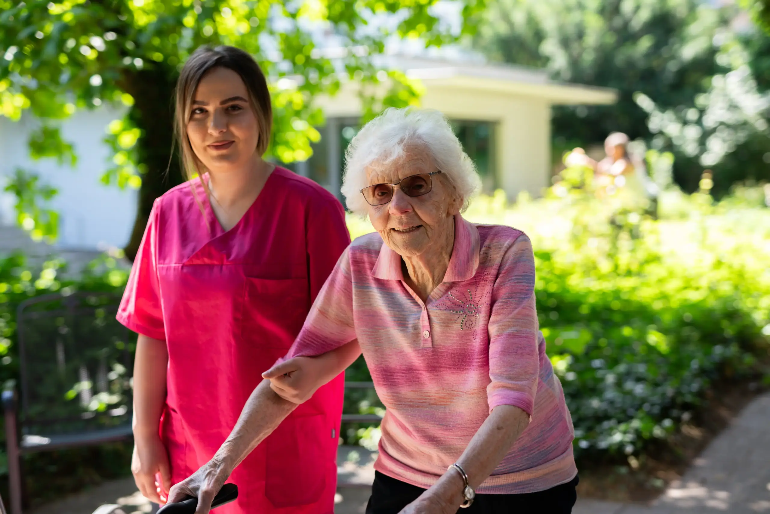Eine jüngere Frau und eine ältere Frau stehen nebeneinander im Garten des Friedensheims. Die ältere Frau hat ihre Hände an einem Rollator, die jüngere Frau trägt rote Pflegekleidung und hat sich bei ihr untergehakt. Sie lächeln in die Kamera.