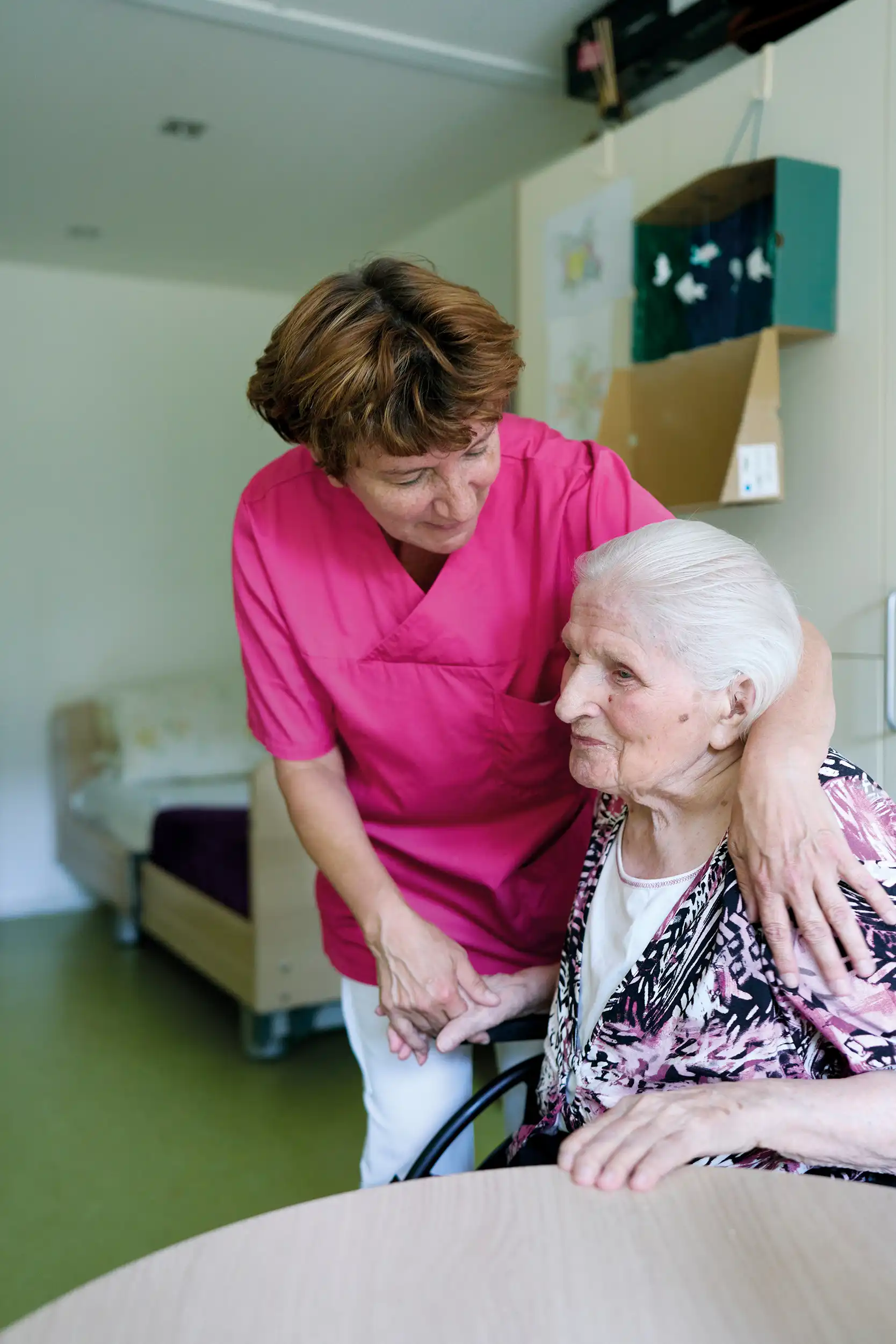 Eine ältere Frau sitzt an einem Tisch. Eine andere Frau in roter Pflegekleidung steht neben ihr und legt den Arm um sie. Im Hintergrund sieht man ein Krankenbett.