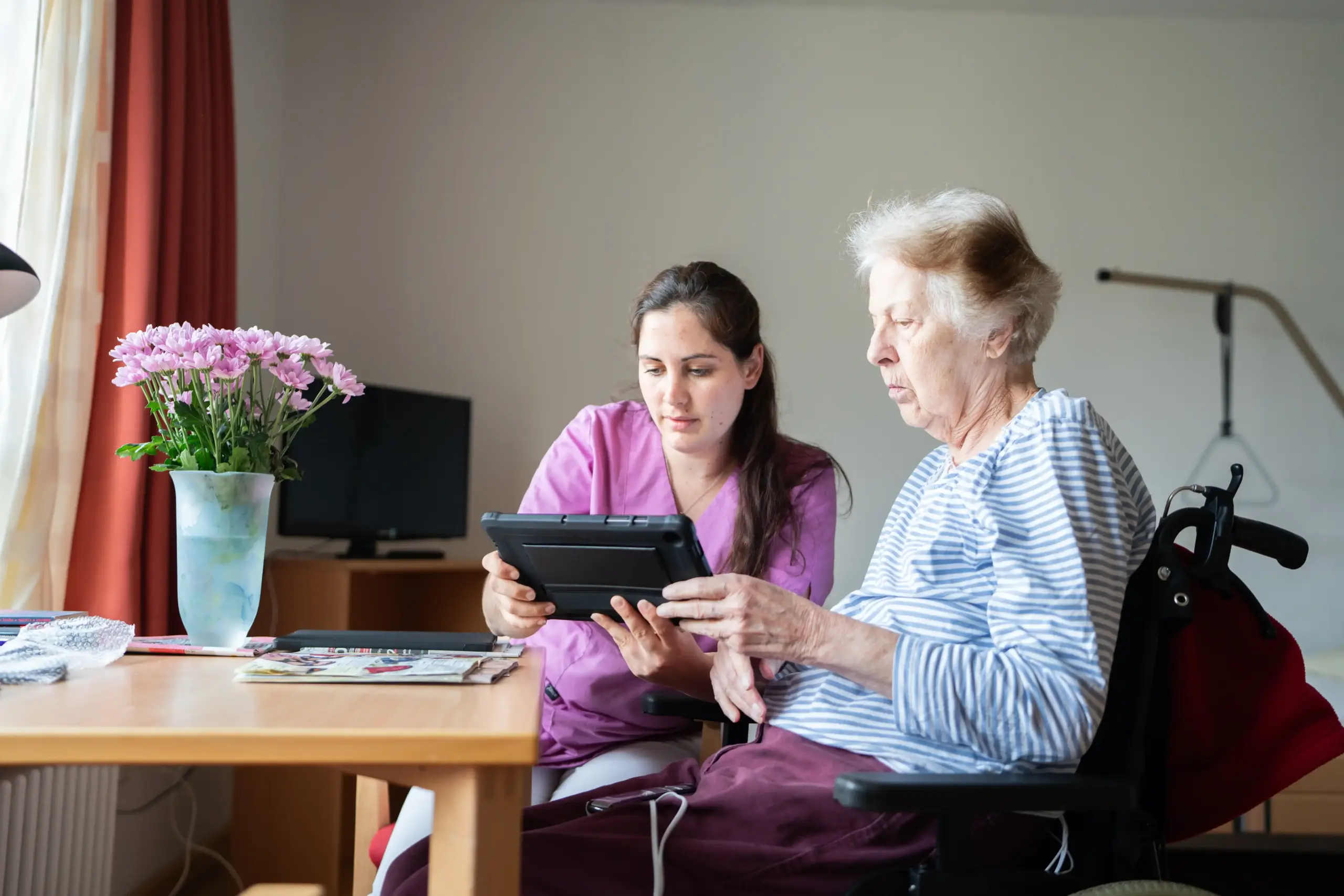Eine ältere Frau und eine jüngere Frau schauen gemeinsam auf ein Tablet. Die ältere Frau sitzt in einem Rollstuhl. Die jüngere Frau trägt violette Pflegekleidung. Im Hintergrund stehen ein Fernseher und ein Bett.