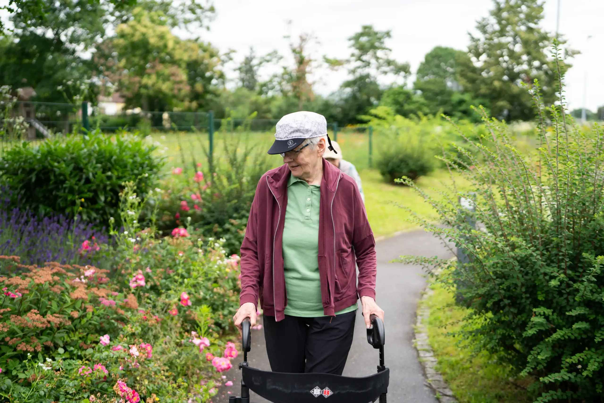Quelle: BLV Zwei Frau spazieren hintereinander mit ihren Rollatoren durch den Garten des Hauses Karlsruher Weg. Sie betrachten die Blumen neben dem Weg.