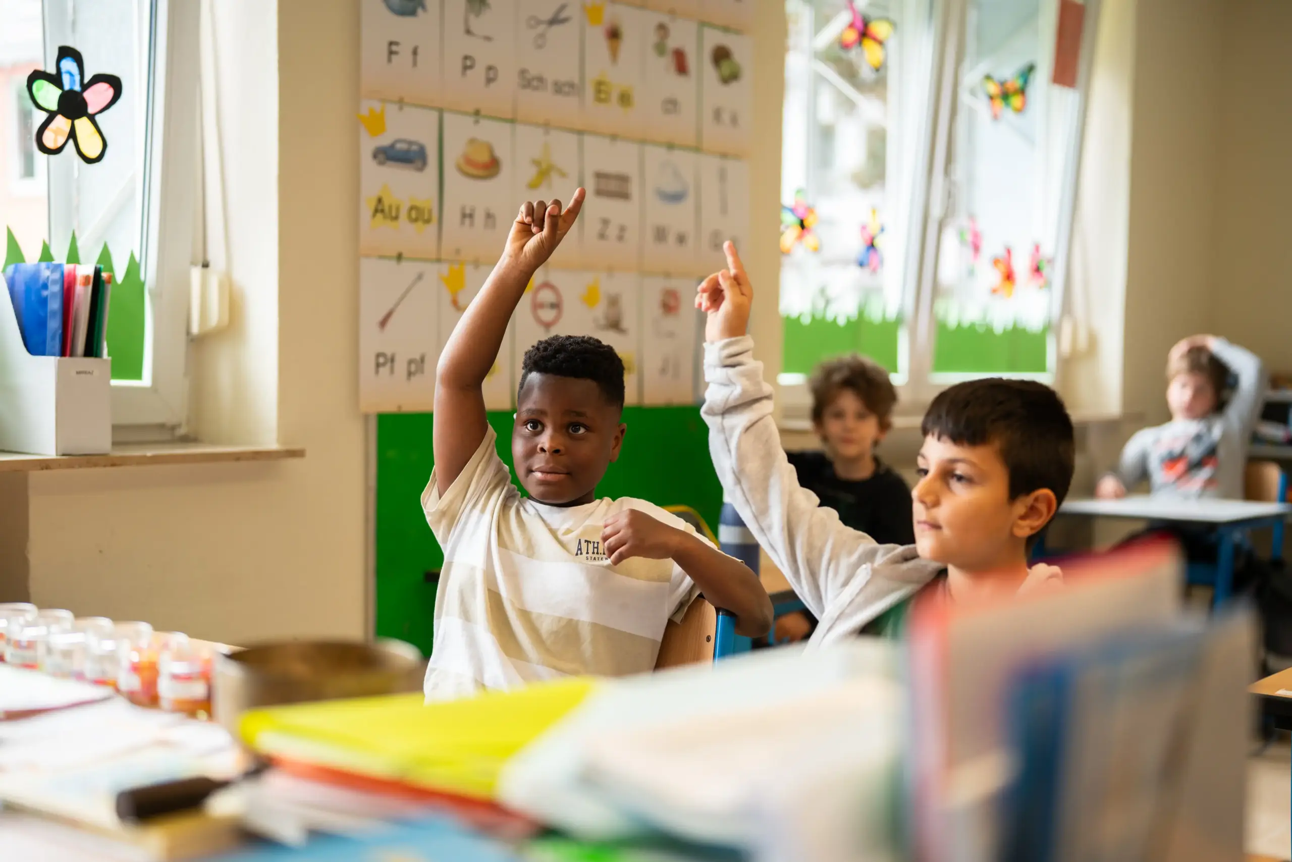 Vier Schüler sitzen an Schultischen. Die vorderen beiden heben die Hand, um sich zu melden.