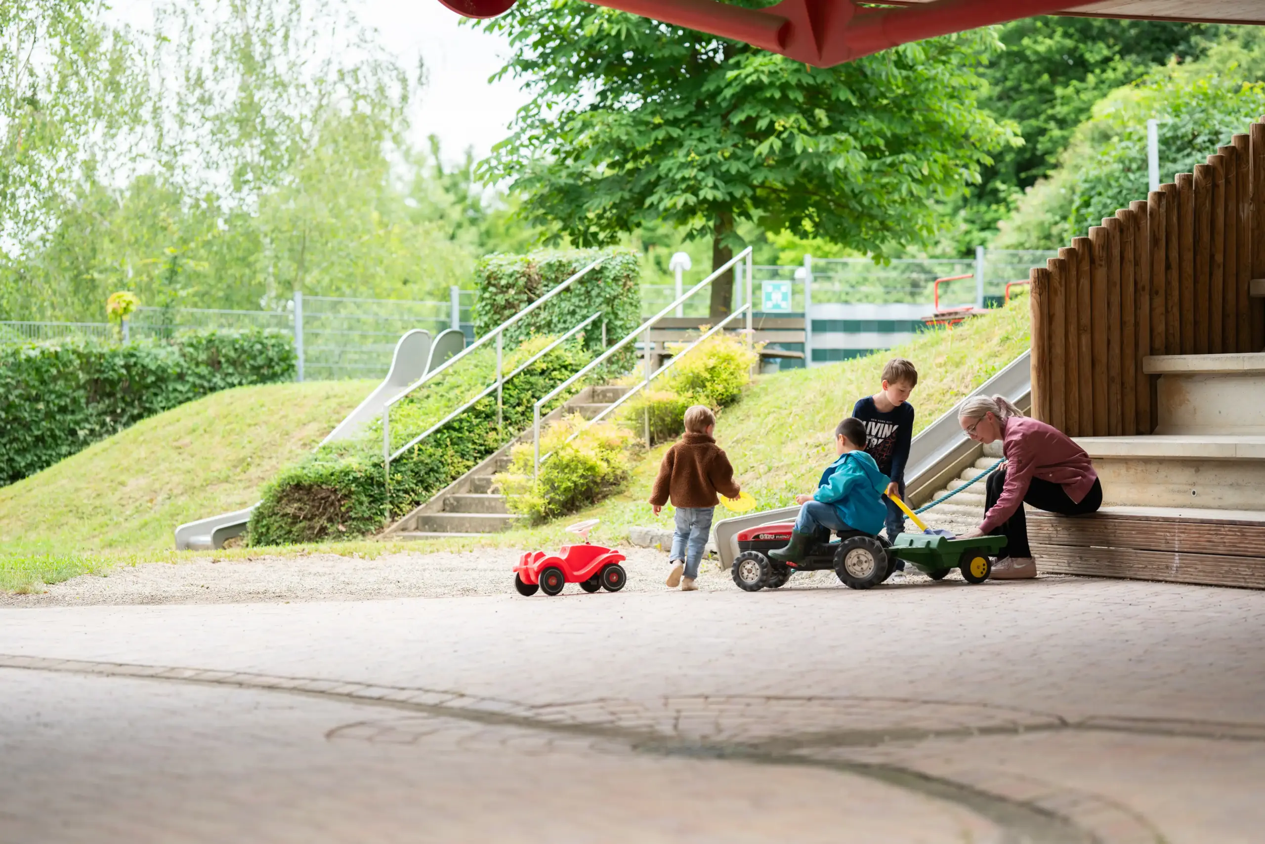 Quelle: BLV Drei Kinder spielen miteinander mit Spielfahrzeugen: einem Bobbycar und einem Traktor. Sie schaufeln Sand in den Anhänger des Traktors. Eine junge Frau sitzt daneben und hilft mit.