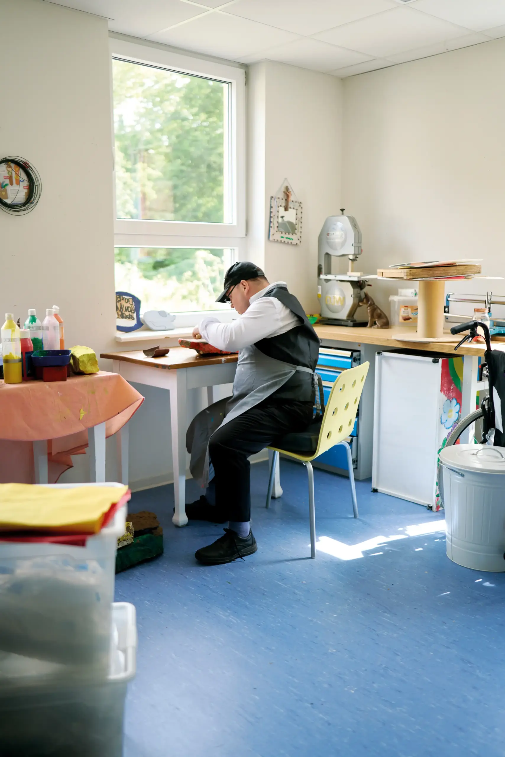 Ein Mann sitzt an einem Tisch und arbeitet handwerklich. Er hat eine graue Schürze um und eine schwarze Mütze auf. Neben im stehen bunte Farbtuben, auf der anderen Seite diverse Bastelgegenstände.