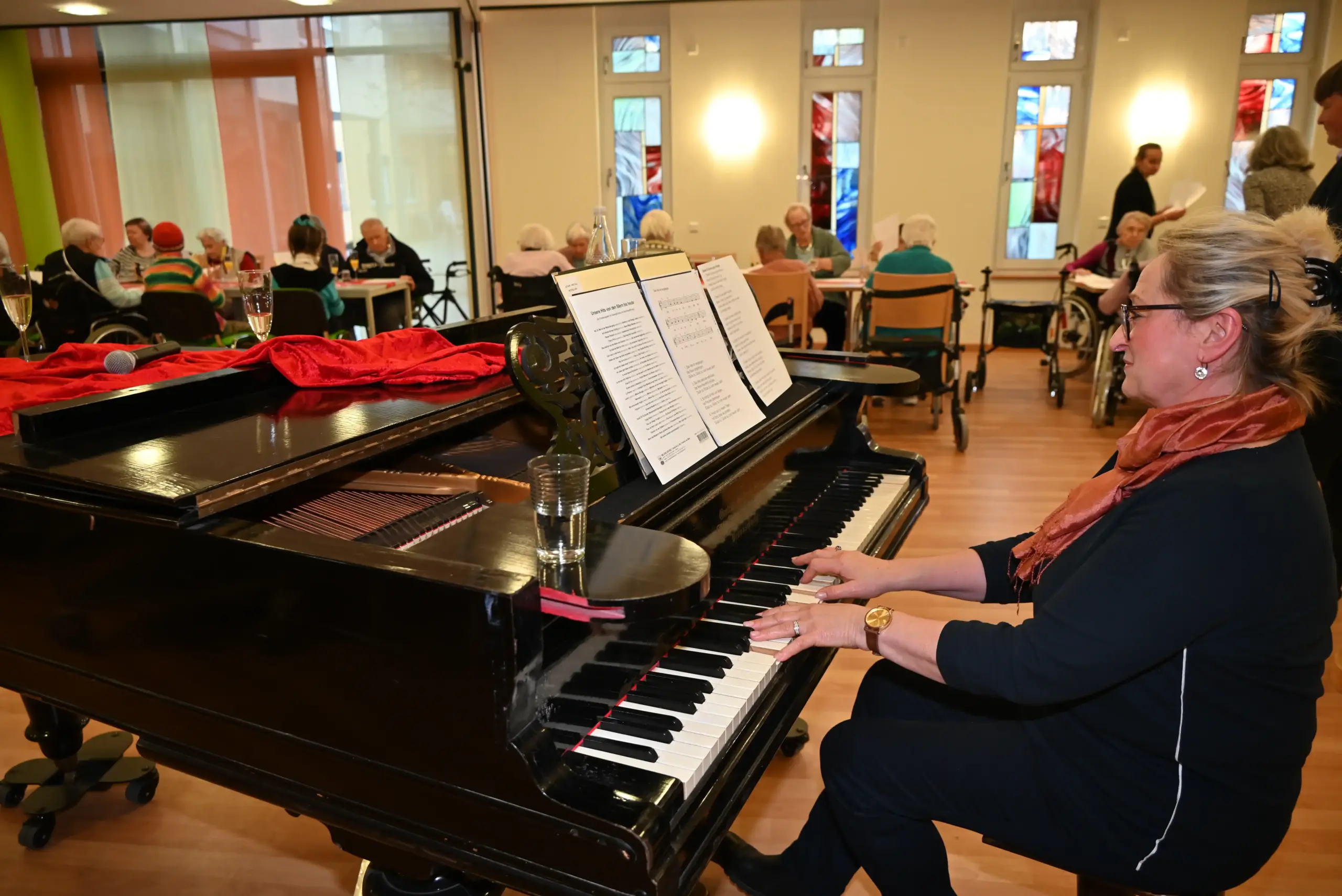 Eine Frau spielt auf einem Flügel Klavier. Im Hintergrund sitzen einige ältere Menschen an kleinen Tischen zusammen. Manche von ihnen haben Sekt auf dem Tisch stehen.