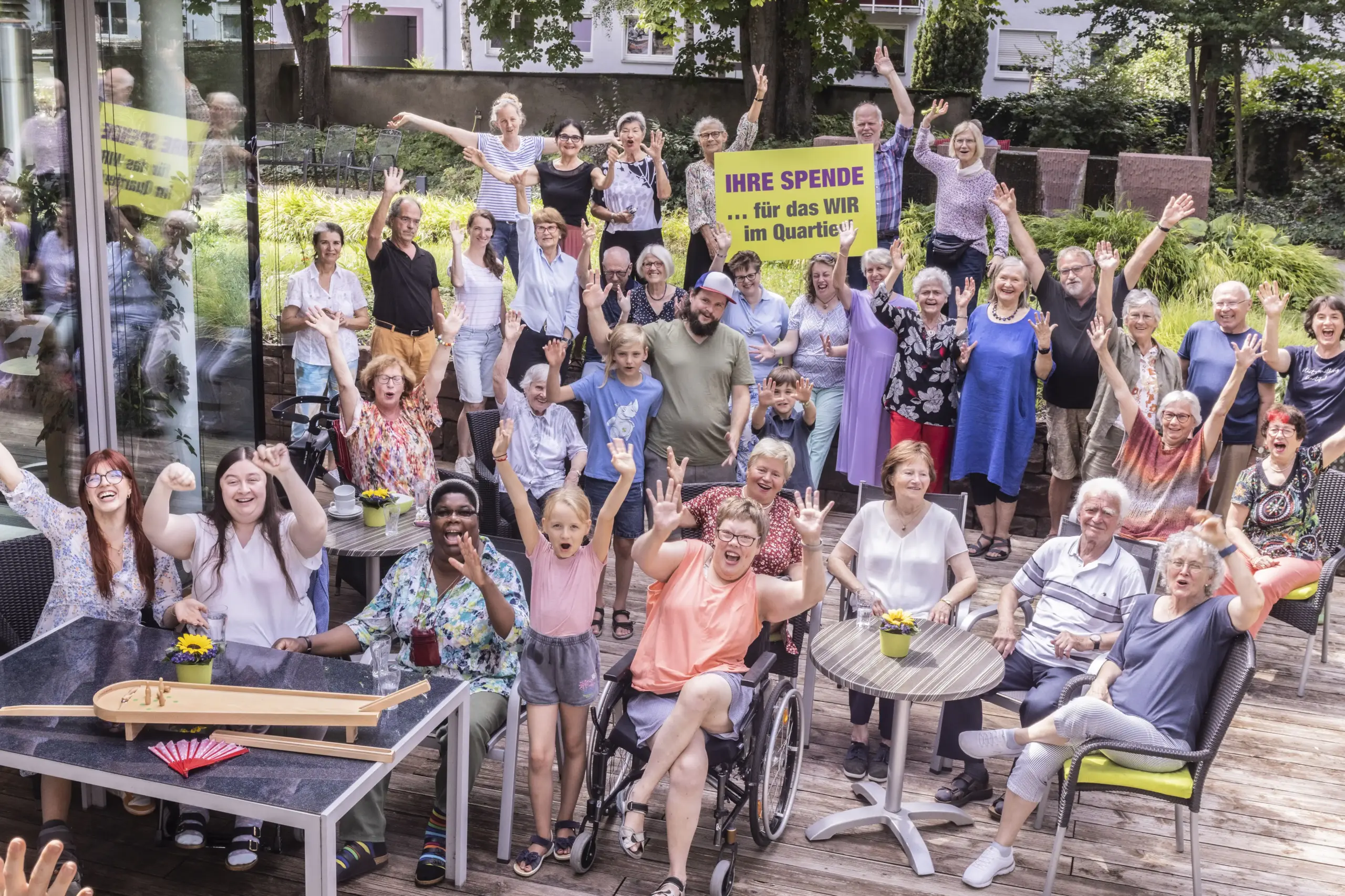Eine große Gruppe Menschen sitzt und steht im Garten des Friedensheims. Sie halten ein Schuld mit der Aufschrift "Ihre Spende ... für das Wir im Quartier!" in die Höhe und winken.