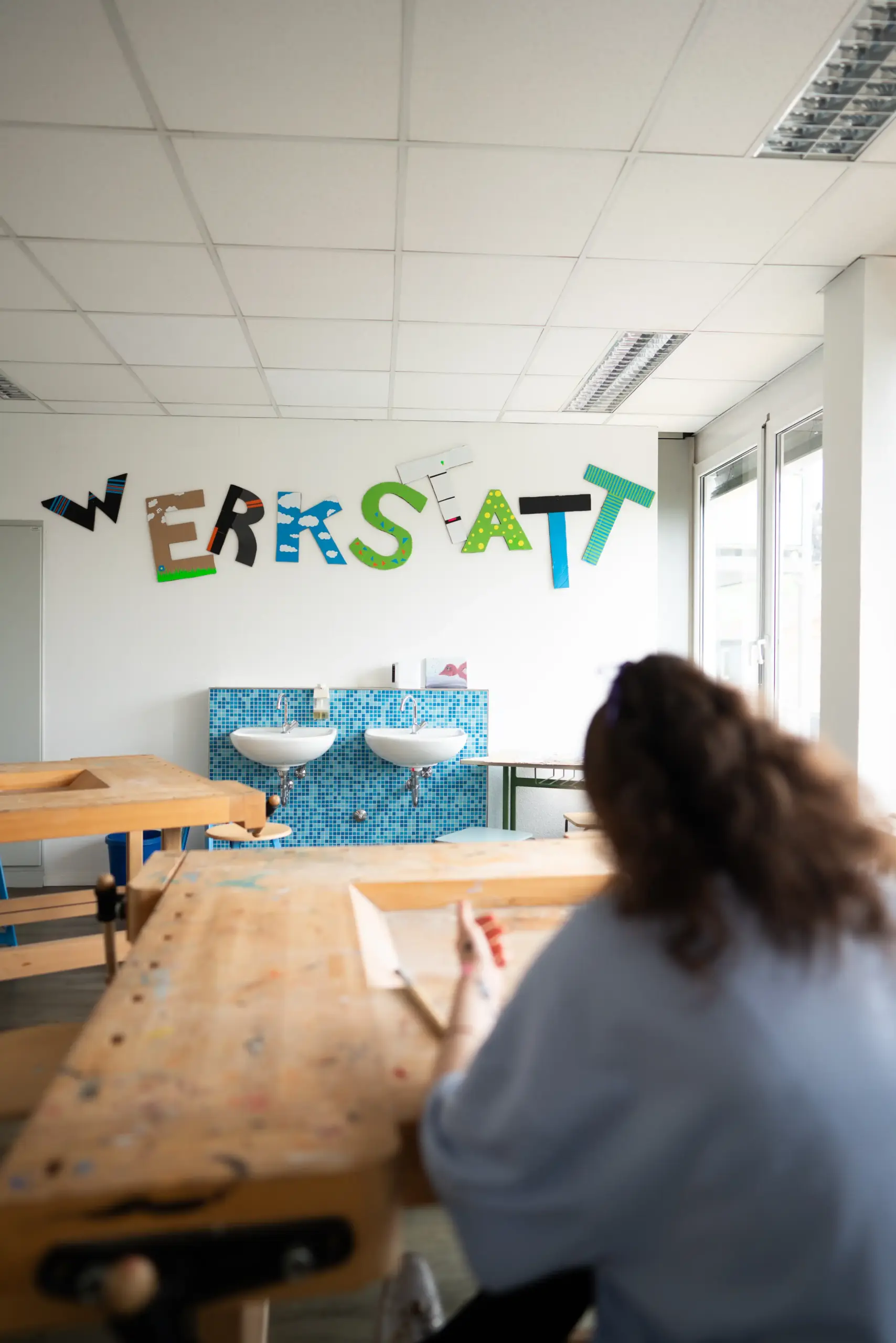 Eine Frau sitzt an einem Tisch und liest. Im Hintergrund steht an der Wand in großen bunten Buchstaben 