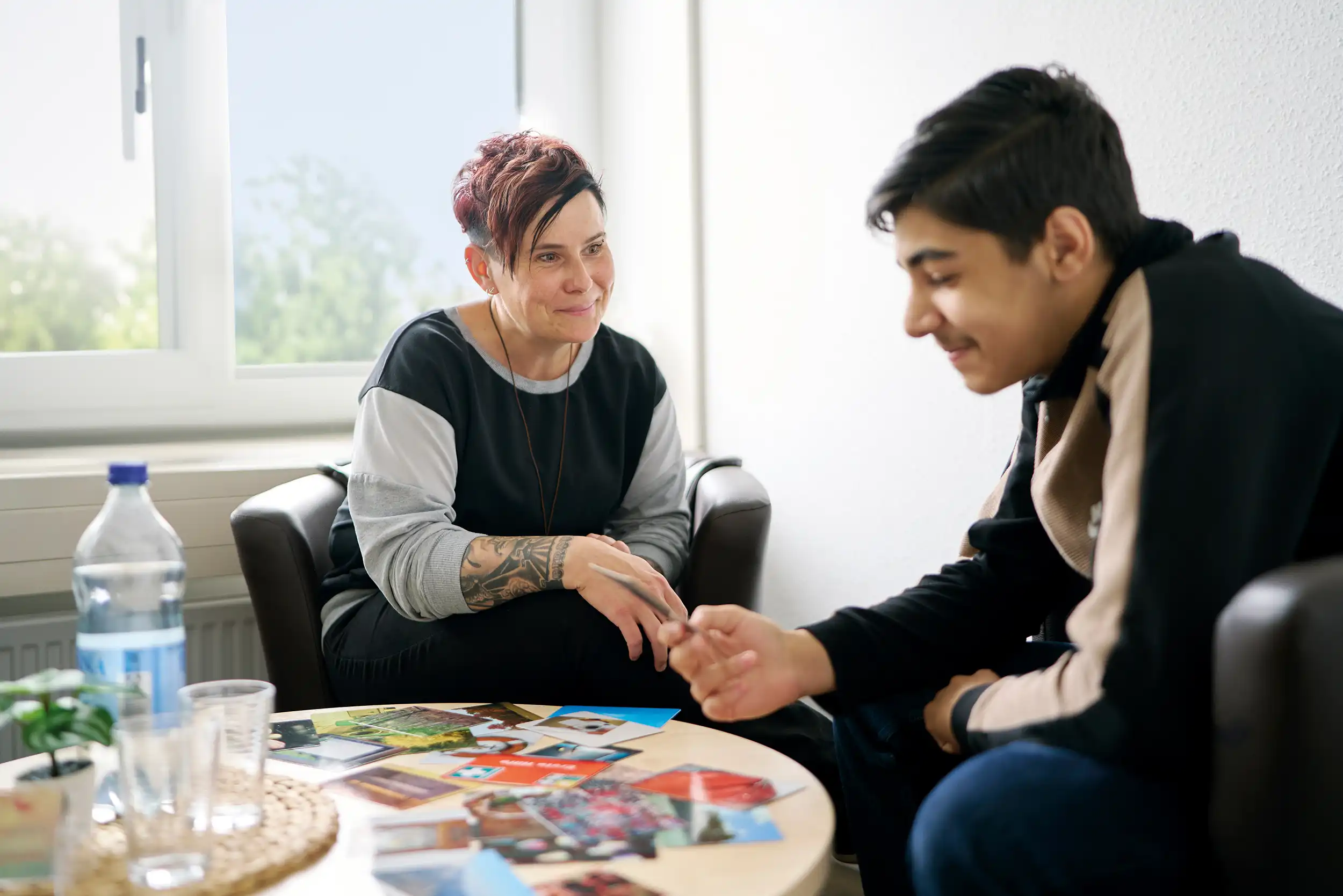 Eine Frau und ein Jugendlicher sitzen auf Stuhlen und unterhalten sich. Auf dem Tisch vor ihnen liegen verschiedene bunte Karten. Beide lachen.