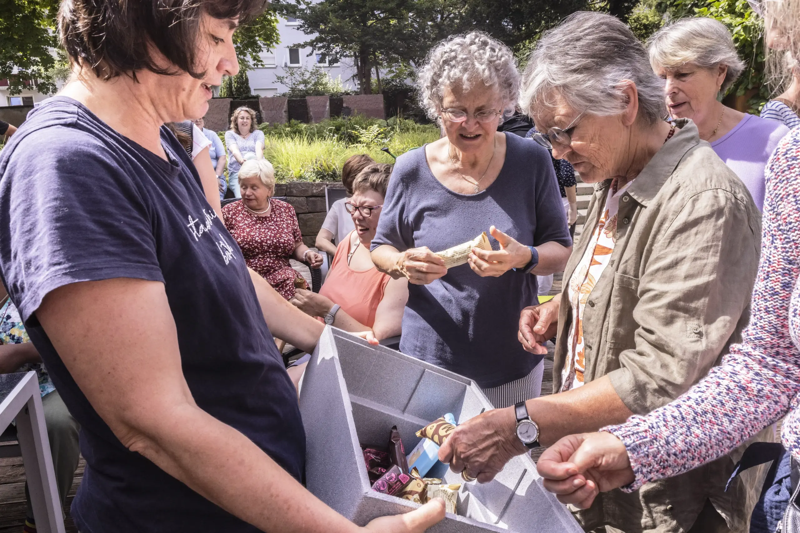 Quartiersmitarbeiterin Monika Scheytt teilt Eis an eine Gruppe älterer Frauen aus. Im Hintergrund sitzen weitere Personen auf Stühlen zusammen.