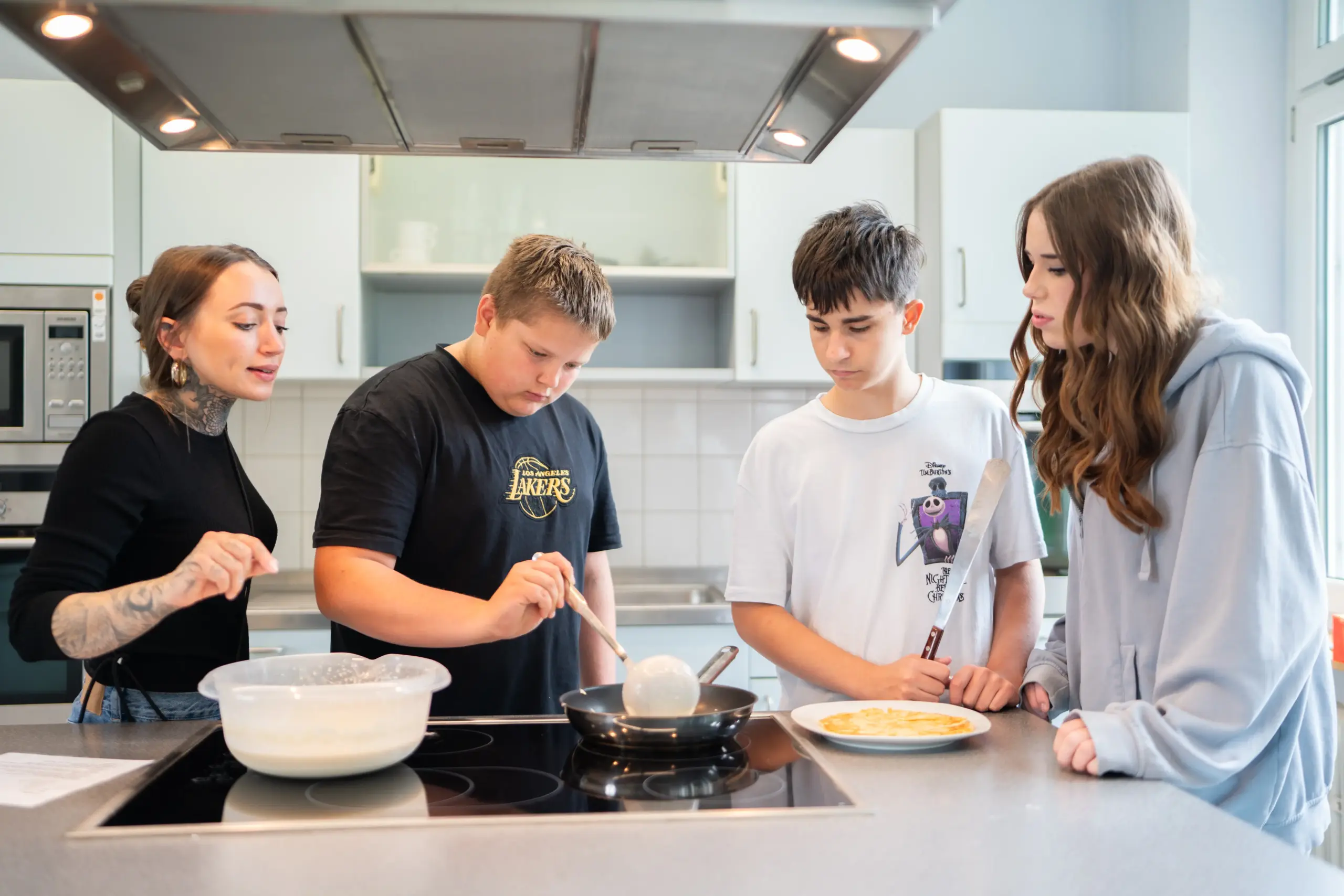 Eine Frau und drei Jugendliche stehen in einer Großküche am Herd. Einer der Jungen schöpft mit einer Kelle Teig für einen Pfannenkuchen in die Pfanne. Die anderen schauen zu.