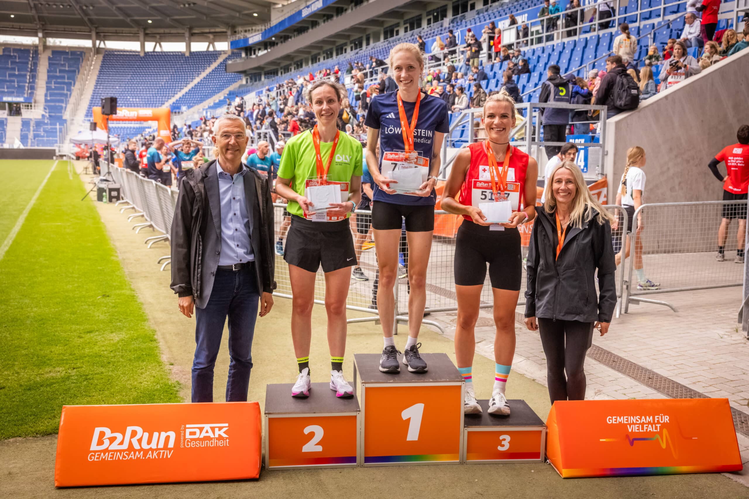 Auf einem Siegertreppchen stehen drei Frauen in Sportkleidung und mit Laufschuhen. Sie haben Medaillen um den Hals, halten ihre Preise in den Händen und lächeln. Links neben dem Podest ein Mann, der Bürgermeister Karlsruhes. Rechts neben dem Podest steht eine Frau. Im Hintergrund kann man die Tribüne eines Stadions erkennen. Hinter einer Absperrung kommen weitere Läuferinnen und Läufer ins Ziel.