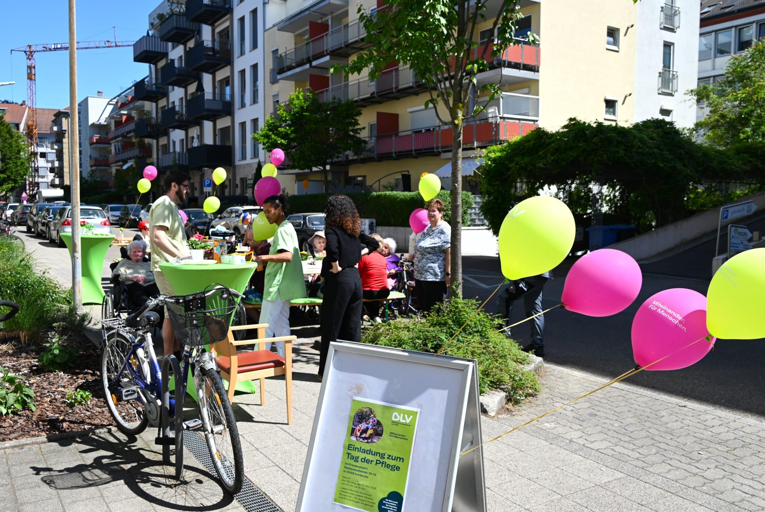 Mehrere Tische und Bänke stehen auf der Straße vor dem Friedensheim. An den Tischen sitzen einige ältere Personen. Manche im Rollstuhl. Die Umgebung ist mit grünen und violetten Luftballons geschmückt.