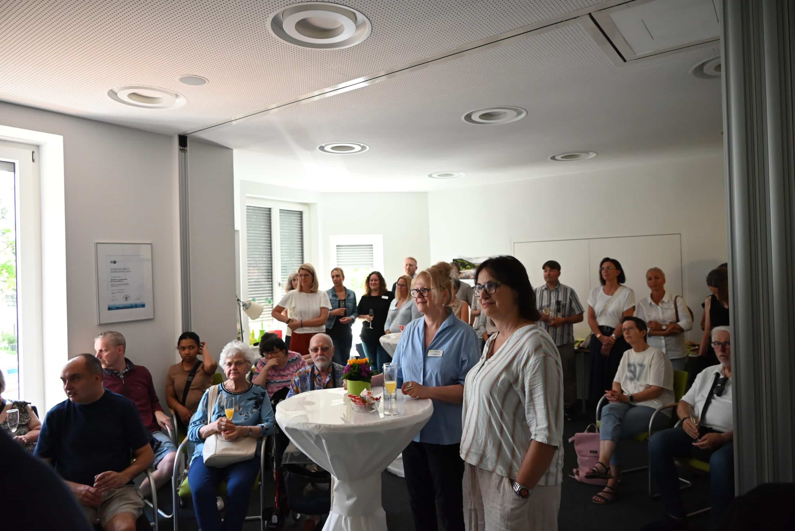 Eine Vielzahl an Personen hören konzentriert zu. Einige sitzen auf Stühlen. Die meisten stehen im Hintergrund zusammen. Im Vordergrund stehen zwei Frauen an einem Stehtisch. Viele der Menschen haben ein Glas Sekt in der Hand.