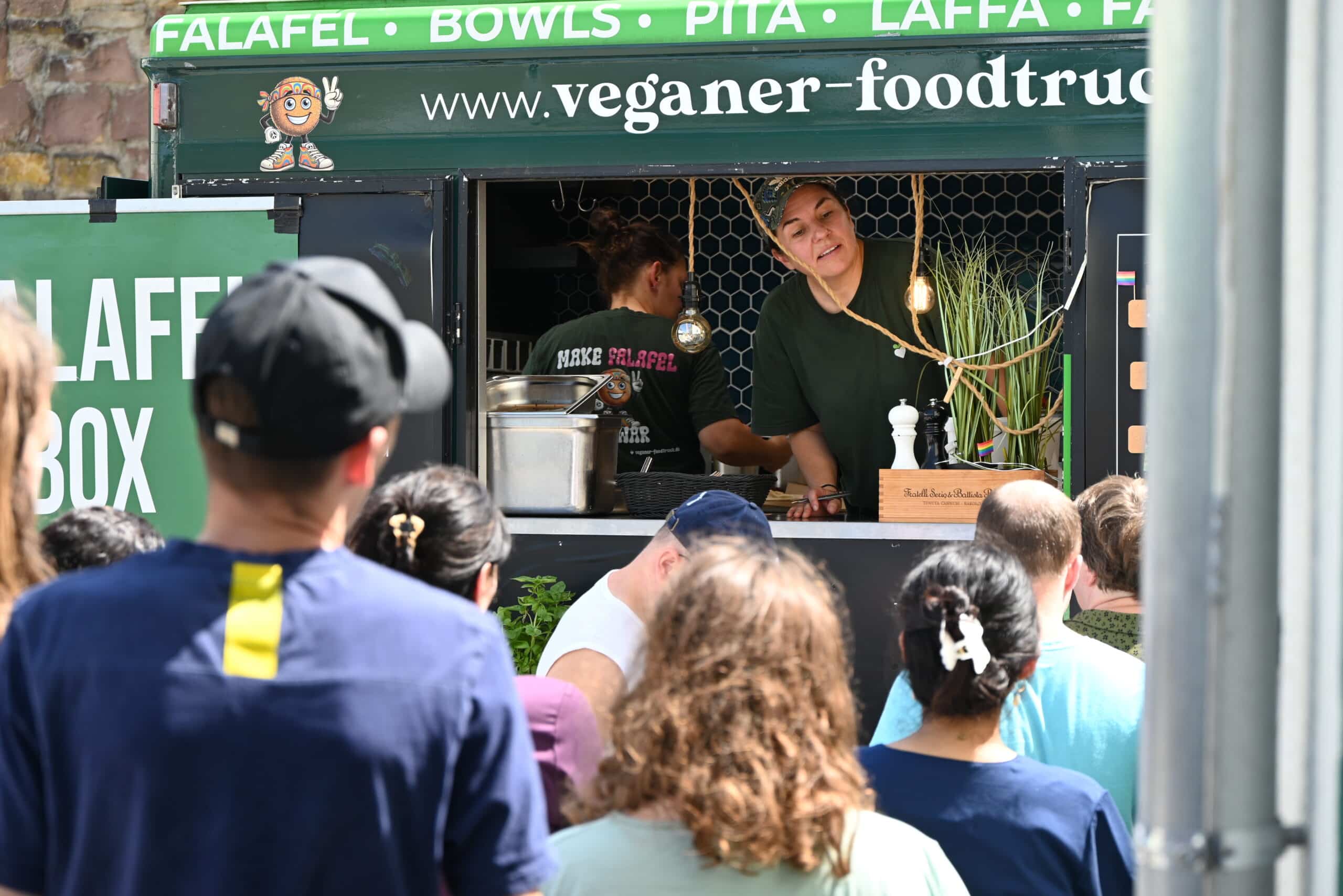 Eine Frau schaut fragend aus dem Fenster des Foodtrucks. Sie nimmt die neuen Essensbestellungen auf. Viele Menschen warten vor dem Foodtruck.