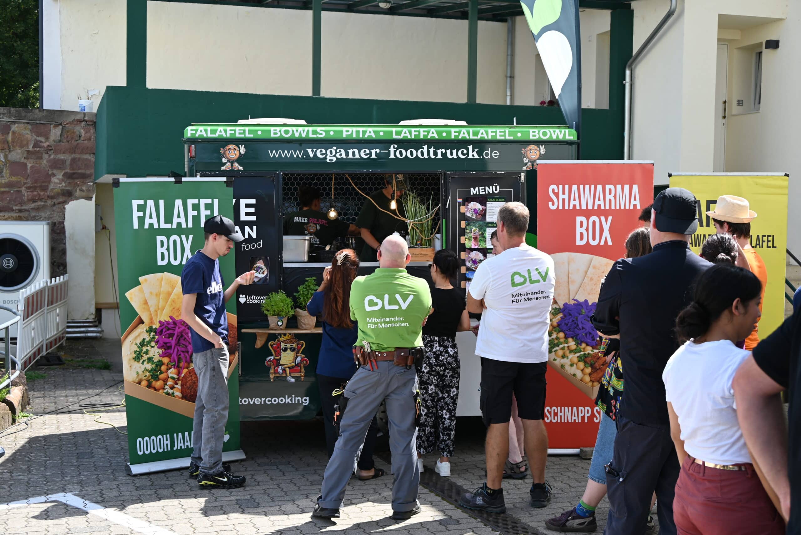 Eine lange Schlange Menschen steht vor einem Foodtruck an und wartet darauf, Essen zu bestellen. Durch ein Fenster sieht man im Innern des Foodtrucks zwei Frauen, wie sie Essen zubereiten. In der Gruppe vor dem Foodtruck sind auch zwei Mitarbeiter des BLV - einer im grünen, der andere im weißen T-Shirt.