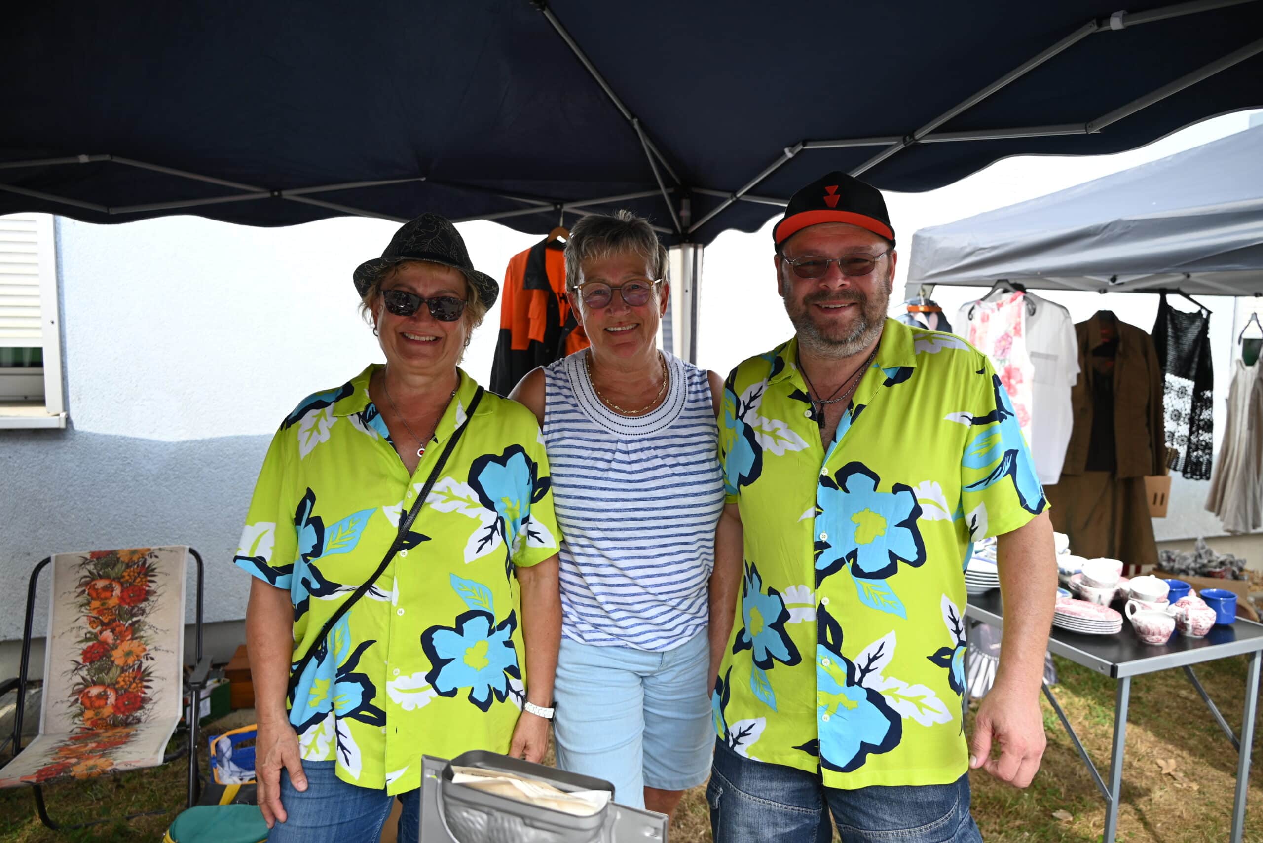 Drei Personen stehen unter einem dunkelblauen Pavillon. Zwei Personen tragen ein hellgrünes Hawaii-Hemd mit großen hellblauen Blüte darauf.