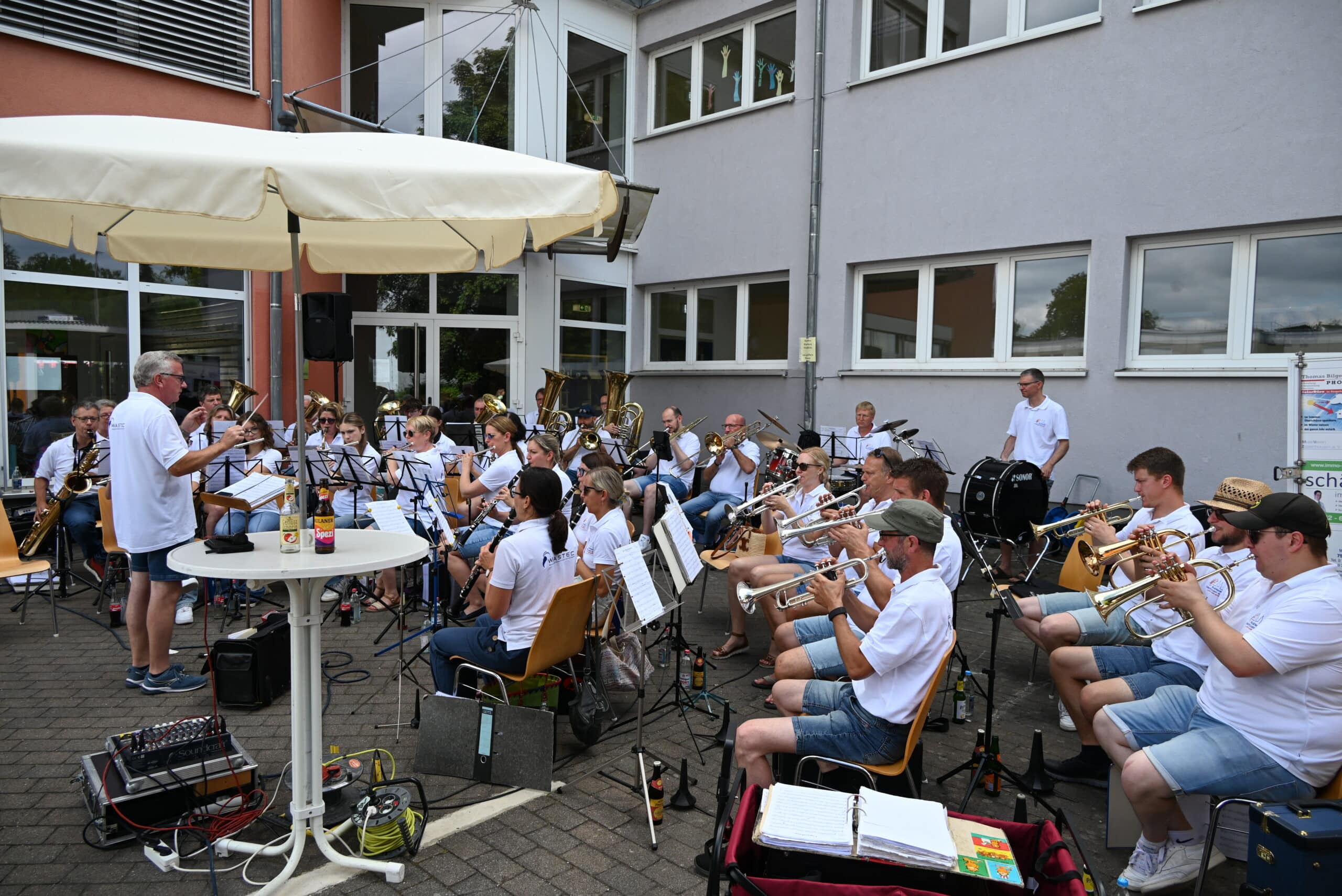 Die Musiker des Musikvereins Zaisenhausen sitzen auf Stühlen vor dem Hauptgebäude der Hohbergschule und spielen ihre Instrumente: Trompeten, Posaunen, Tuba, Klarinetten, Querflöte, Saxophon und viel mehr. Ein Mann steht vor dem Orchester und dirigiert. Alle tragen weiße Poloshirts.