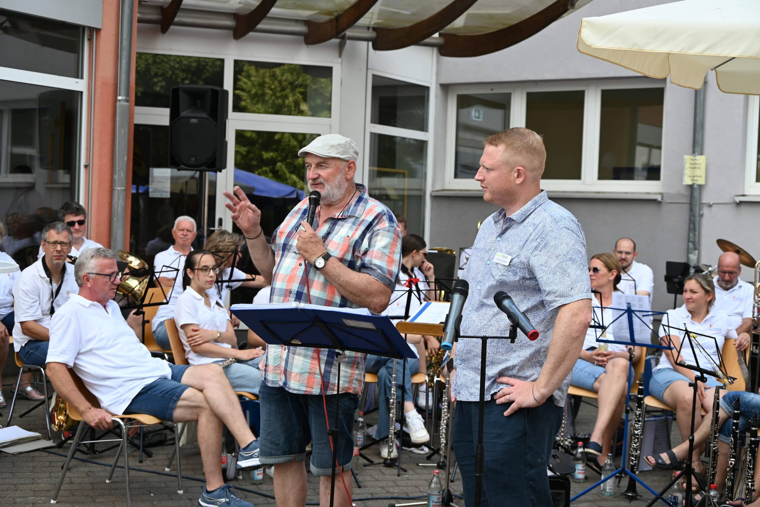 Peter Miebach steht neben Christoph Röckinger und hält eine Rede. Er trägt eine Schiebermütze und ein kariertes Hemd und hält ein Mikrophon in der Hand. Im Hintergrund sitzen die Mitglieder eines Orchesters. Sie haben ihre Musikinstrumente vor sich abgestellt und hören zu.