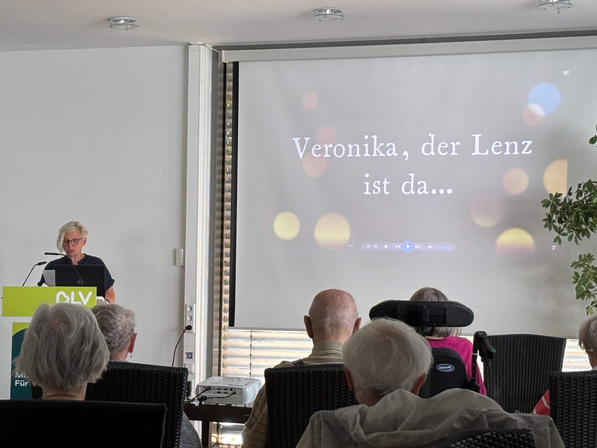 Frau Kammerer steht an einem Pult und spricht. Neben ihr wird mit einem Beamer eine Präsentation auf eine Leinwand gestrahlt. Mehrere Menschen sitzen im Publikum und hören zu.