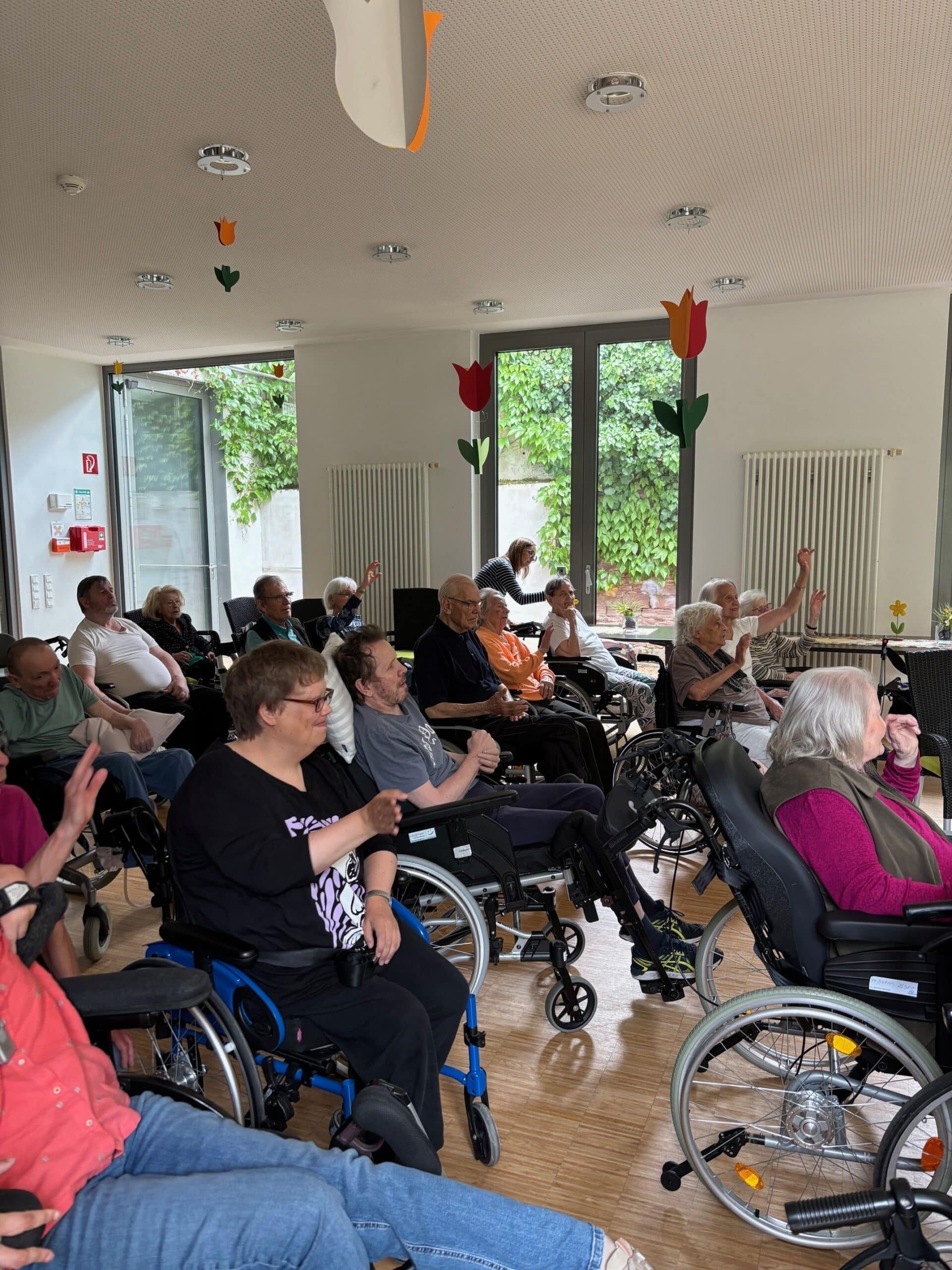 Viele ältere und jüngere Menschen sitzen nebeneinander im Publikum. Die meisten sitzen im Rollstuhl. Sie singen und klatschen.