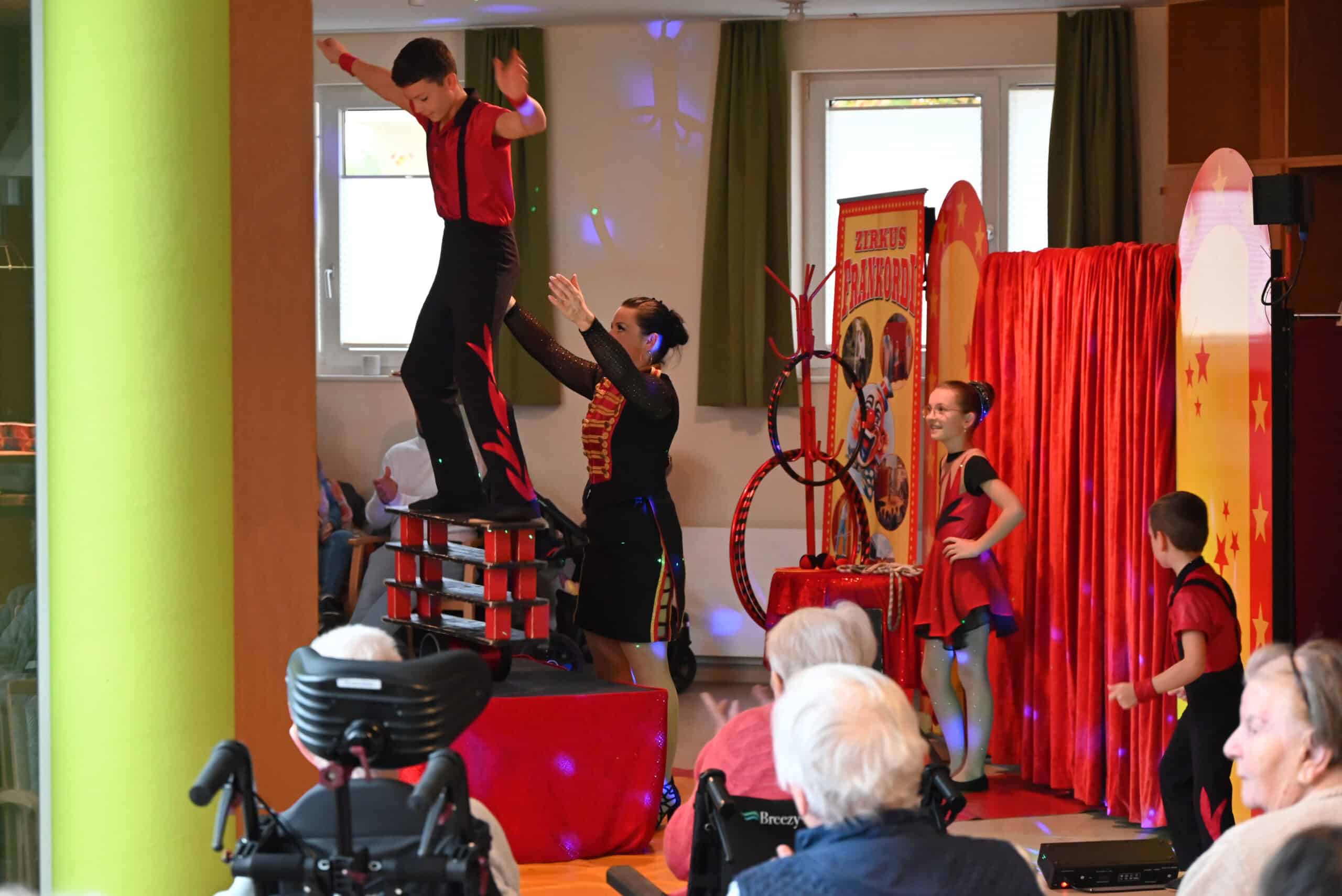 Ein Junge balanciert auf einer Konstruktion aus mehreren Brettern und einer Rolle auf einem Tisch in der Mitte einer kleinen Zirkusmanege. Eine Frau steht neben ihm und streckt die Arme aus, um ihn abzusichern. Mehrere ältere Menschen verfolgen die Vorstellung gebannt.
