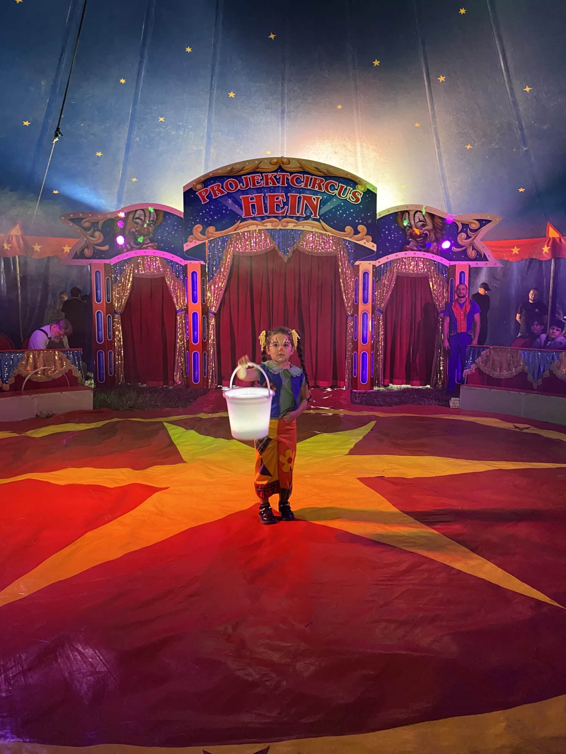 Ein kleines Mädchen steht in der Mitte einer Zirkusmanege. Sie ist geschminkt und trägt bunte Clowns-Kleidung. In der Hand hält sie einen großen weißen Eimer, den sie nach vorne streckt.