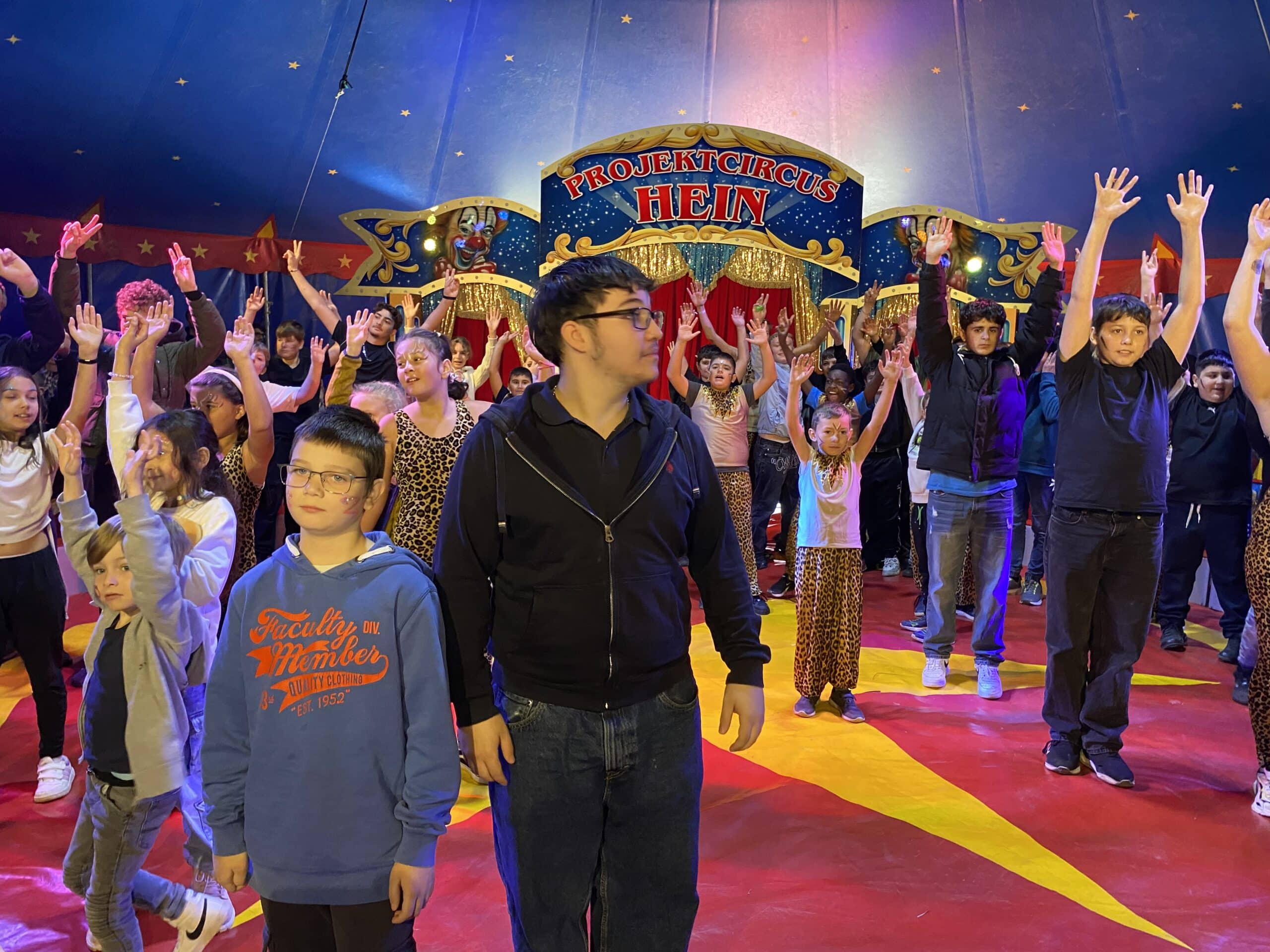 Viele Kinder stehen in der Manege eines Zirkuszelts. Sie lachen und werfen die Arme nach oben.