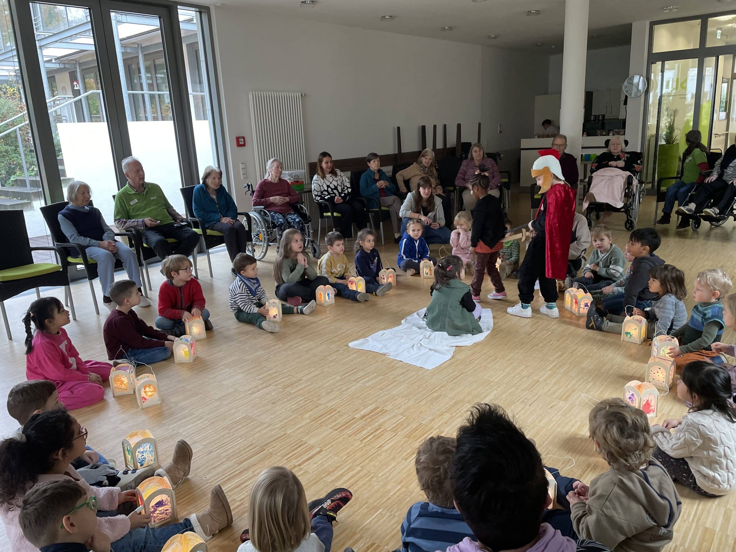 Quelle: BLV Kinder sitzen im Kreis auf dem Boden. Vor ihnen stehen Laternen auf dem Boden. Auf Stühlen sitzen ältere Menschen ebenfalls im Kreis um die Kinder herum.