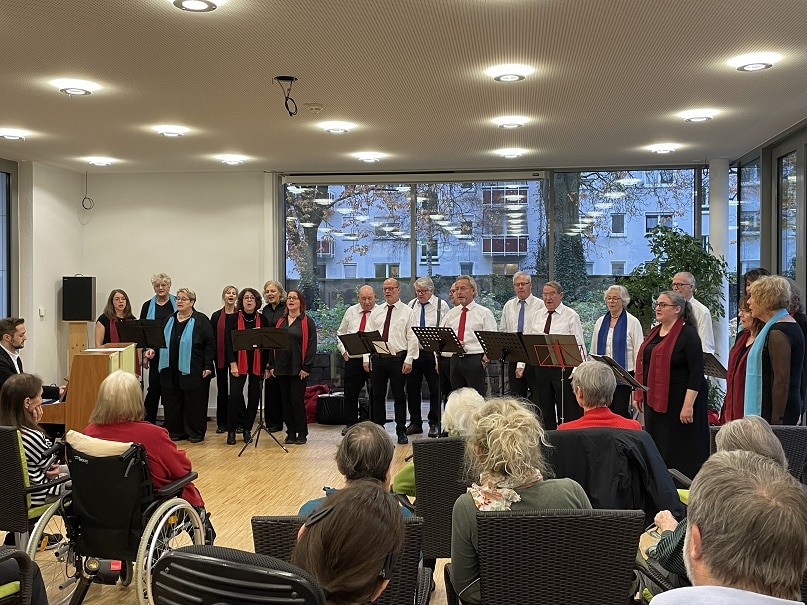 Quelle: BLV Etwa 20 Männer und Frauen stehen im Halbkreis und singen. Neben ihnen steht ein Klavier, auf dem ein Mann spielt. Vor ihnen sitzen mehrere ältere Menschen im Publikum und hören zu. Eine von ihnen sitzt in einem Rollstuhl