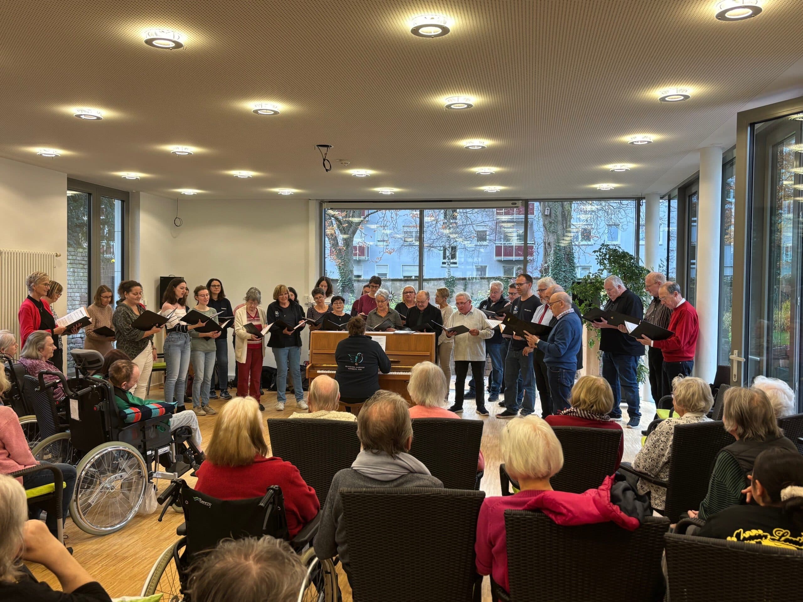 Quelle: BLV Mehrere Menschen stehen im Halbkreis um ein Klavier herum und singen. Auf dem Klavier spielt eine Frau. Vor dem Chor sitzen viele Menschen im Publikum auf Stühlen und höhren zu.