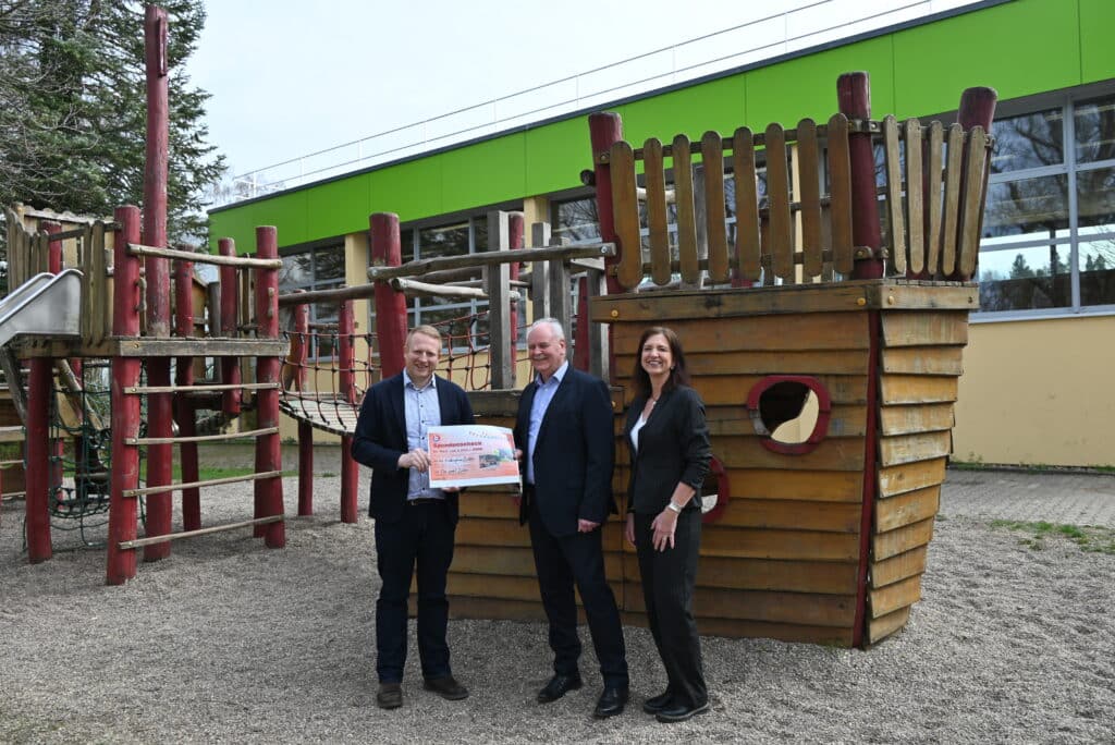 Drei Personen stehen auf einem Spielplatz vor einem Holz-Klettergerüst in Form eines Schiffs und halten einen großen Spendenscheck.