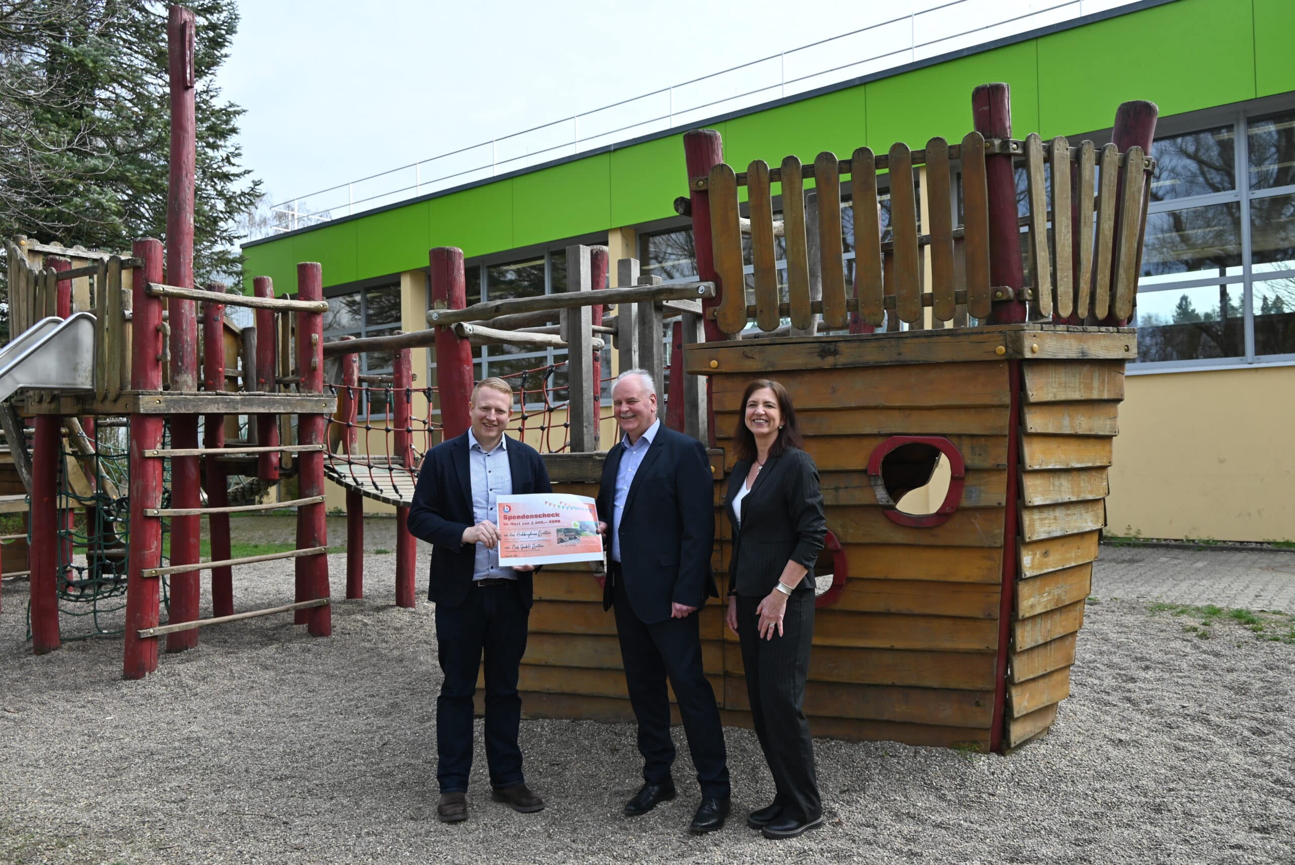 Drei Personen stehen auf einem Spielplatz vor einem Holz-Klettergerüst in Form eines Schiffs und halten einen großen Spendenscheck.