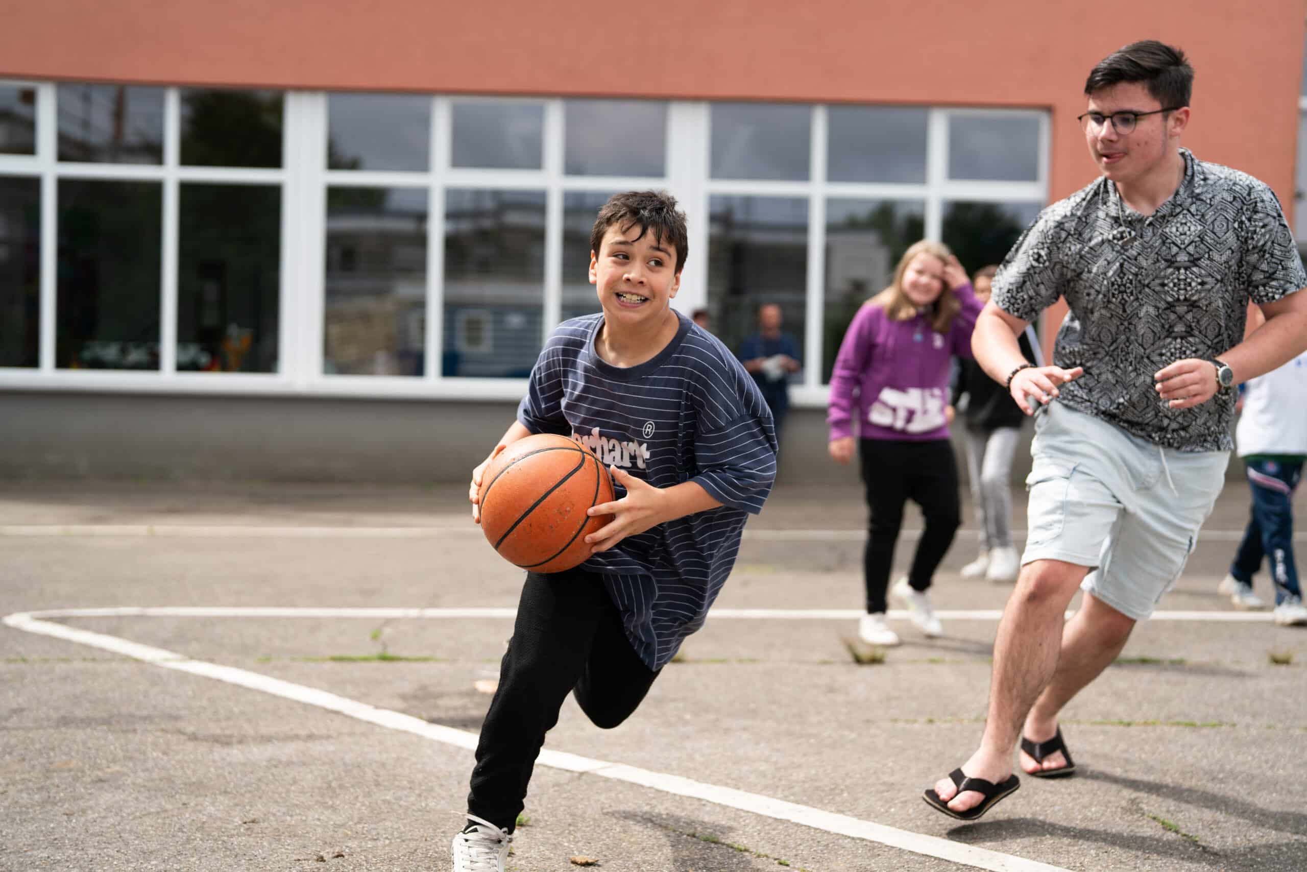 Zwei Jungen rennen, einer der beiden hat einen Basketball in den Händen. Im Hintergrund sind weitere Kinder zu sehen.