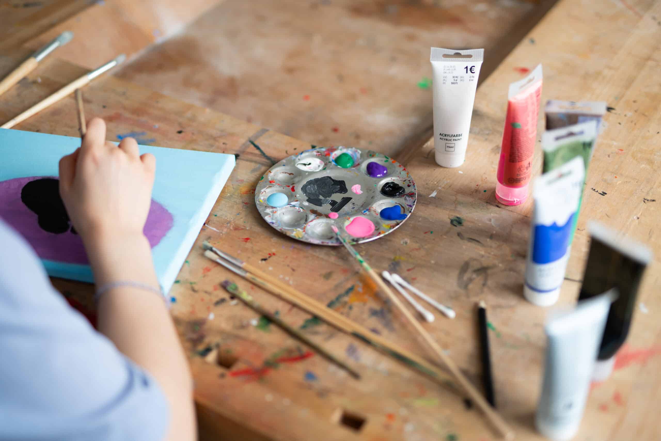 Eine Person mal mit Acrylfarbe. Mehrere Farbtuben stehen auf dem Tisch.