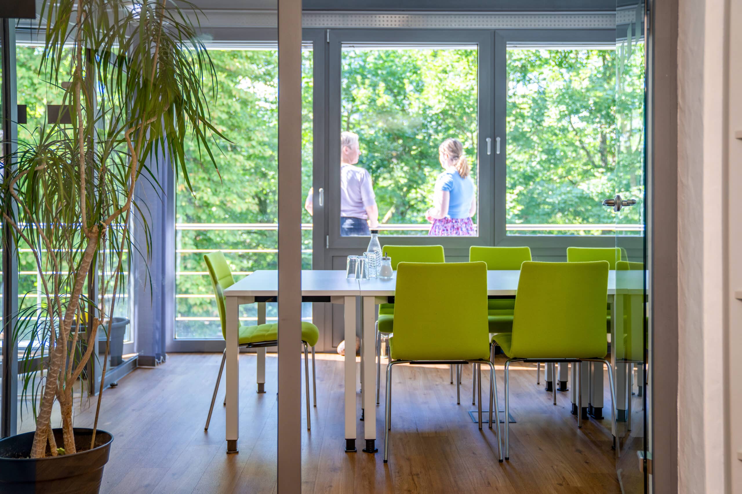 Im Vordergrund steht ein Tisch mit sechs grünen Stühlen. Im Hintergrund stehen zwei Frauen auf einem Balkon und sprechen miteinander.