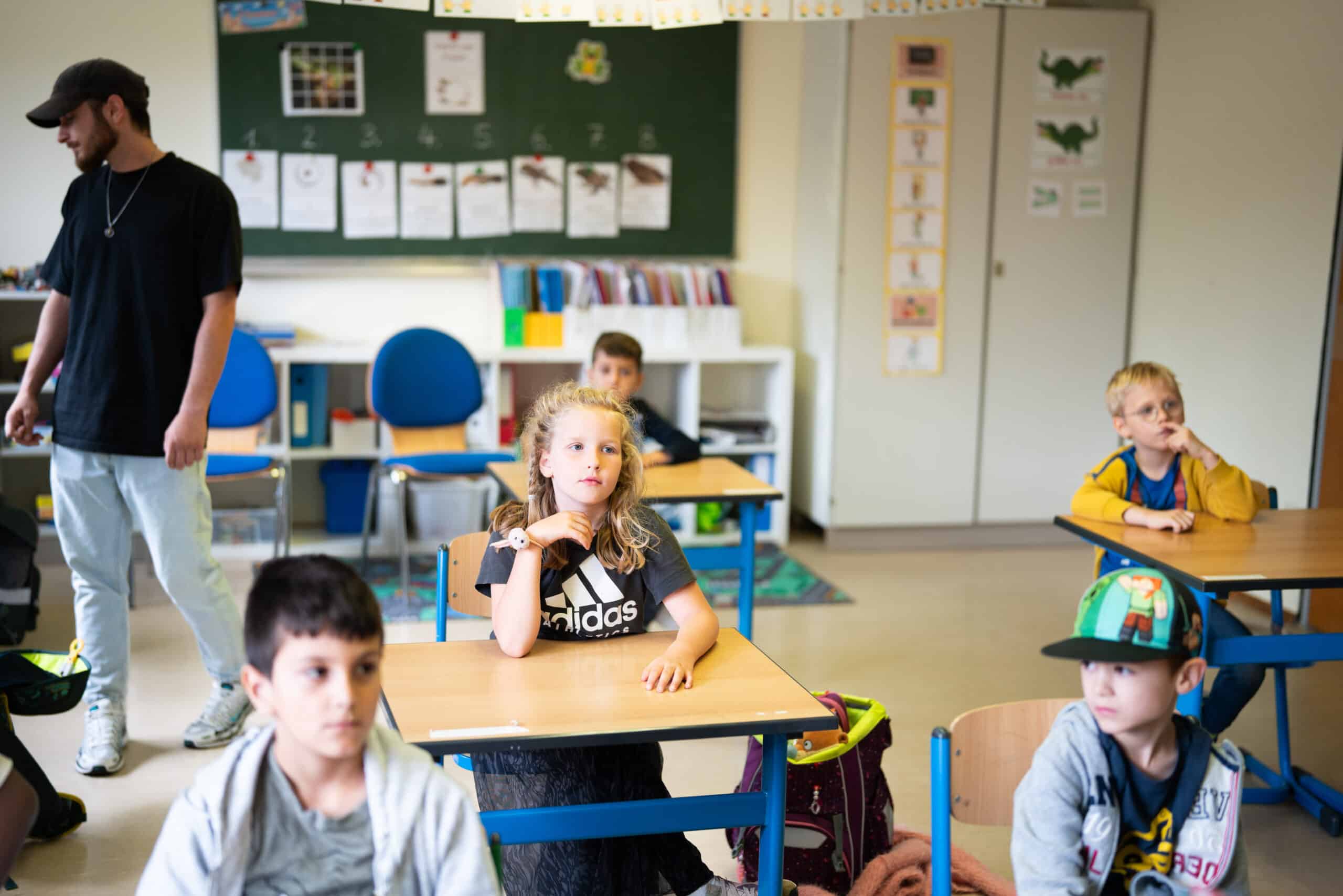 Eine Klassensituation: fünf Kinder sitzen an einzelnen Schultischen und lauschen dem Unterricht. An der Seite steht eine erwachsene Betreuungsperson.