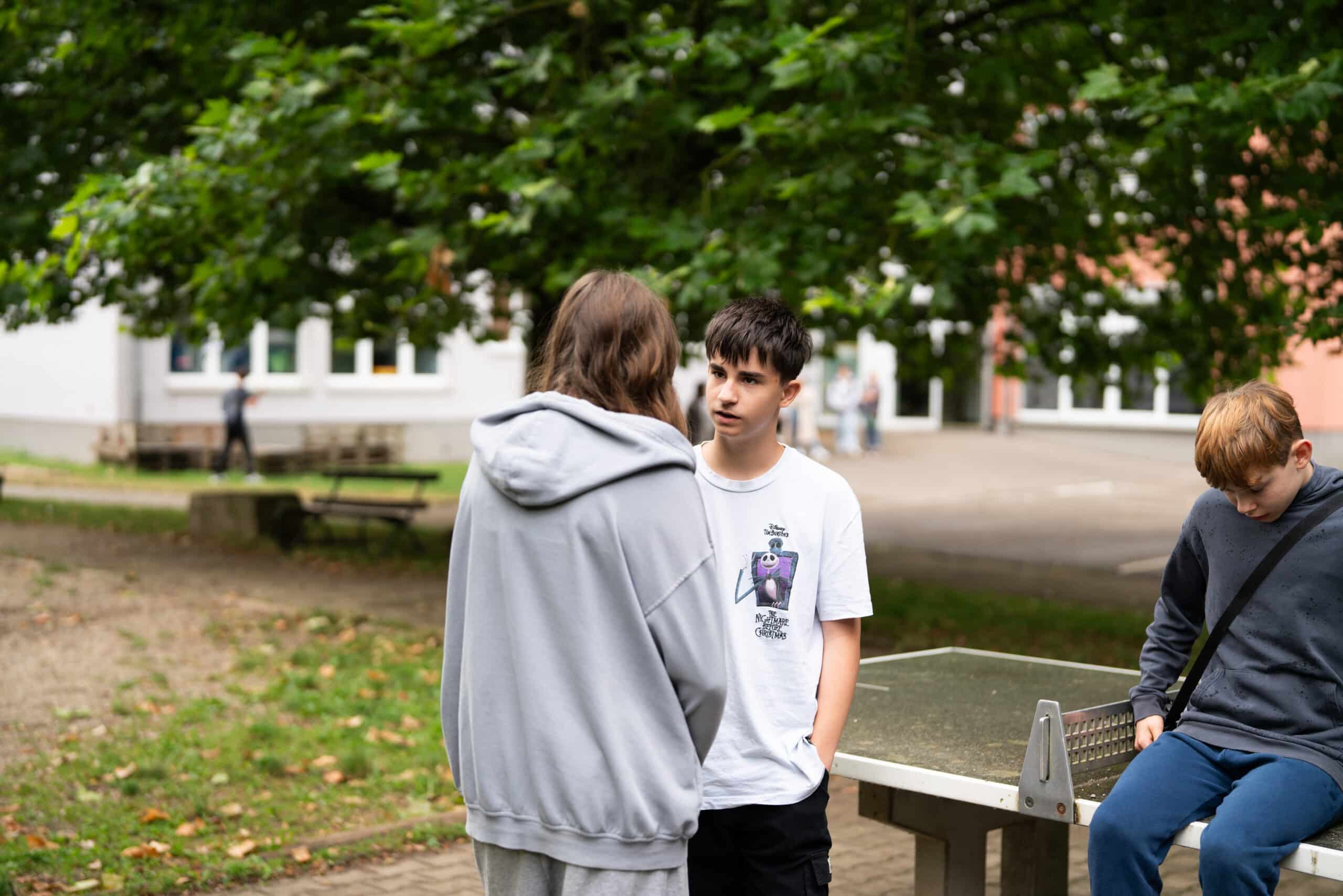 Zwei Jugendliche, ein Junge und ein Mädchen, stehen neben einer Tischtennisplatte und unterhalten sich. Auf der Tischtennisplatte sitzt ein weiterer Junge.