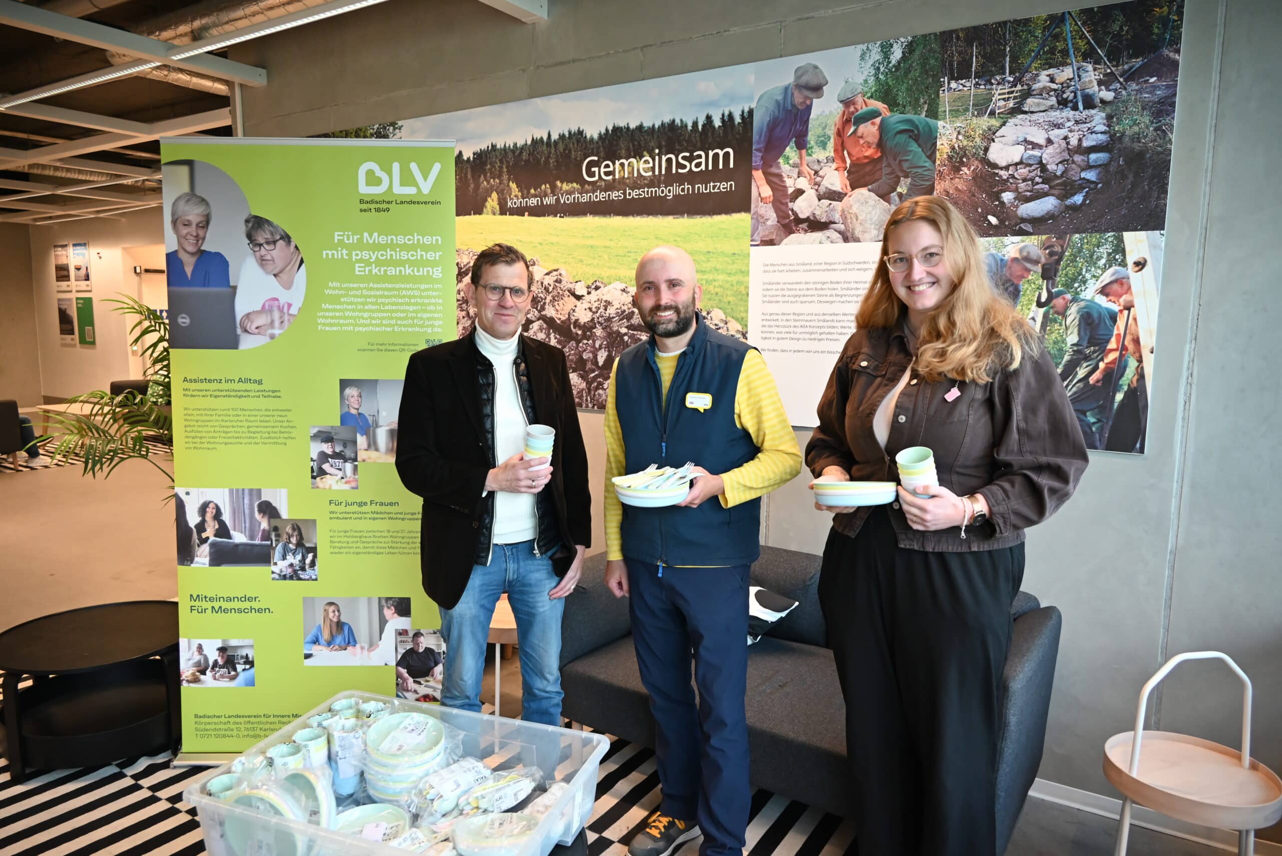 Zwei Männer und eine Frau stehen vor einem großen Plakat von IKEA auf dem steht 