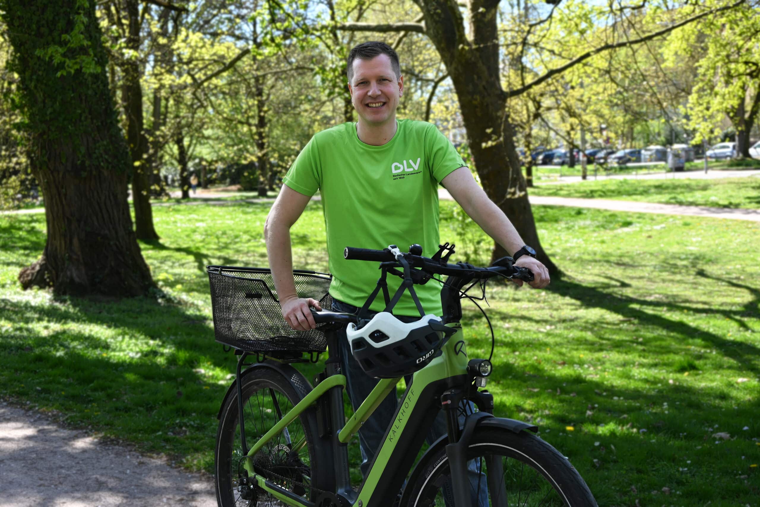 Quelle: BLV Timo Müller steht an seinem grünen Fahrrad vor Bäumen auf einer Wiese und lächelt in die Kamera.