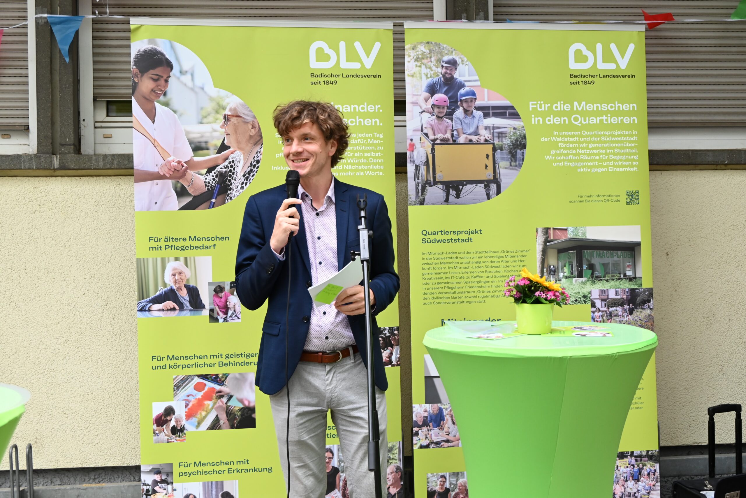 Dr. Raimund Bauer, Leiter der Strategischen Angebotsentwicklung des BLV steht vor zwei grünen Roll-Ups und spricht in ein Mikrofon. Neben ihm steht ein Stehtisch mit grüner Husse und einem Blumengesteck aus Sonnenblumen.