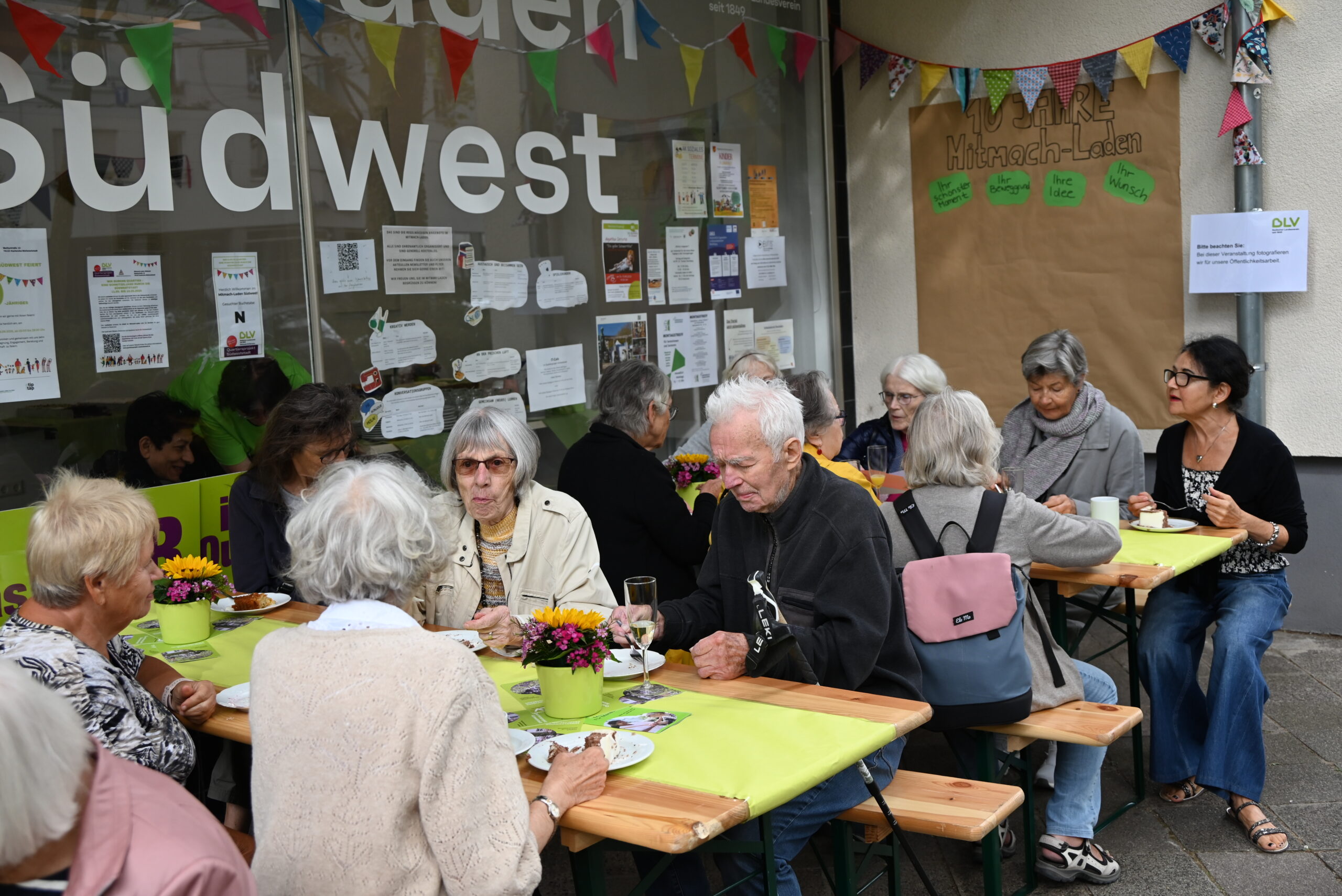 Viele Menschen sitzen vor dem Mitmach-Laden Südwest auf Bierbänken und essen Kuchen. Auf den Tischen stehen bunte Blumensträuße in hellgrünen Töpfen. Im Hintergrund hängt eine bunte Girlande.