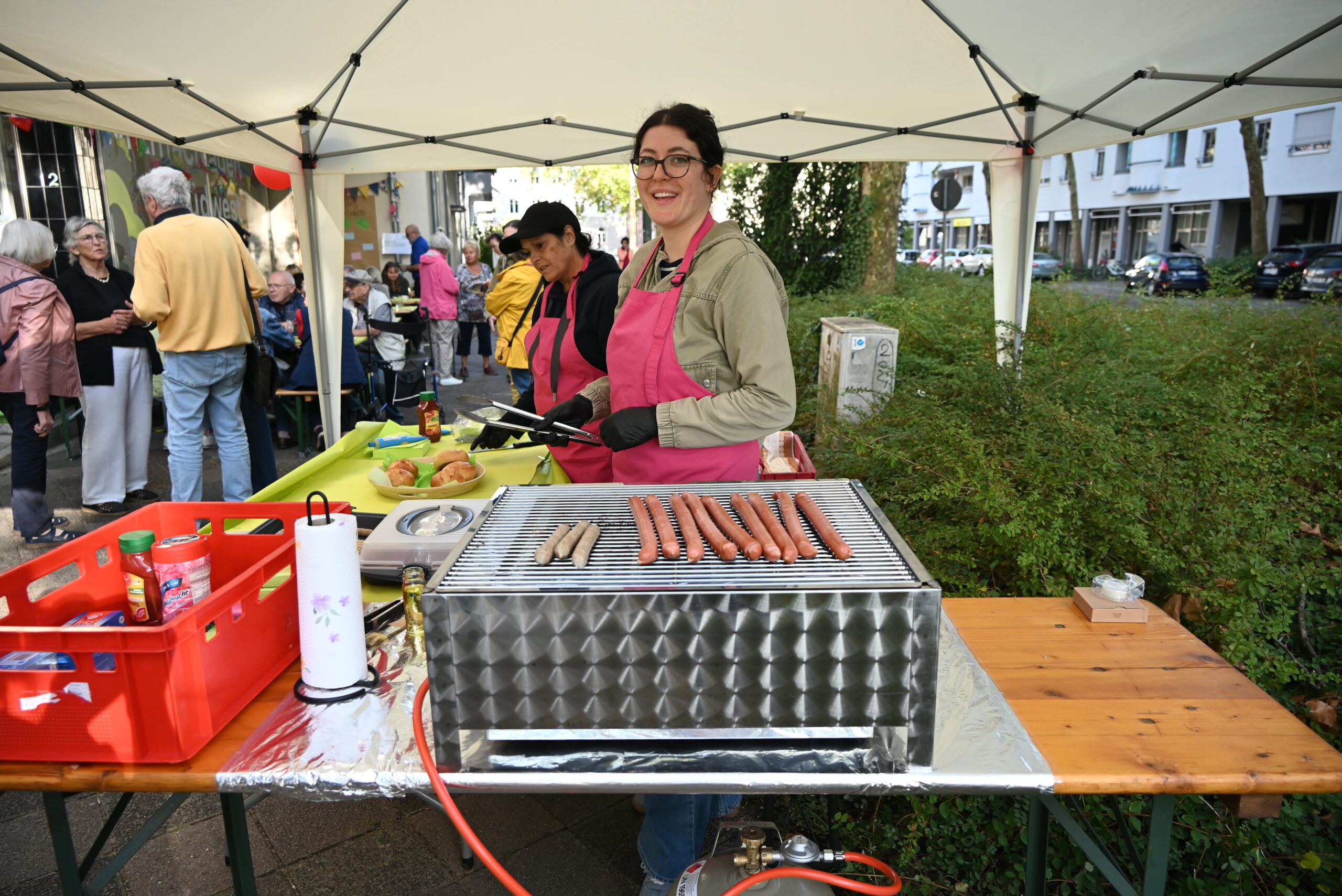 Zwei Frauen stehen am Grill und grillen Bratwürste. Sie tragen rosa Schürzen und lächeln in die Kamera. Im Hintergrund unterhalten sich mehrere Menschen.